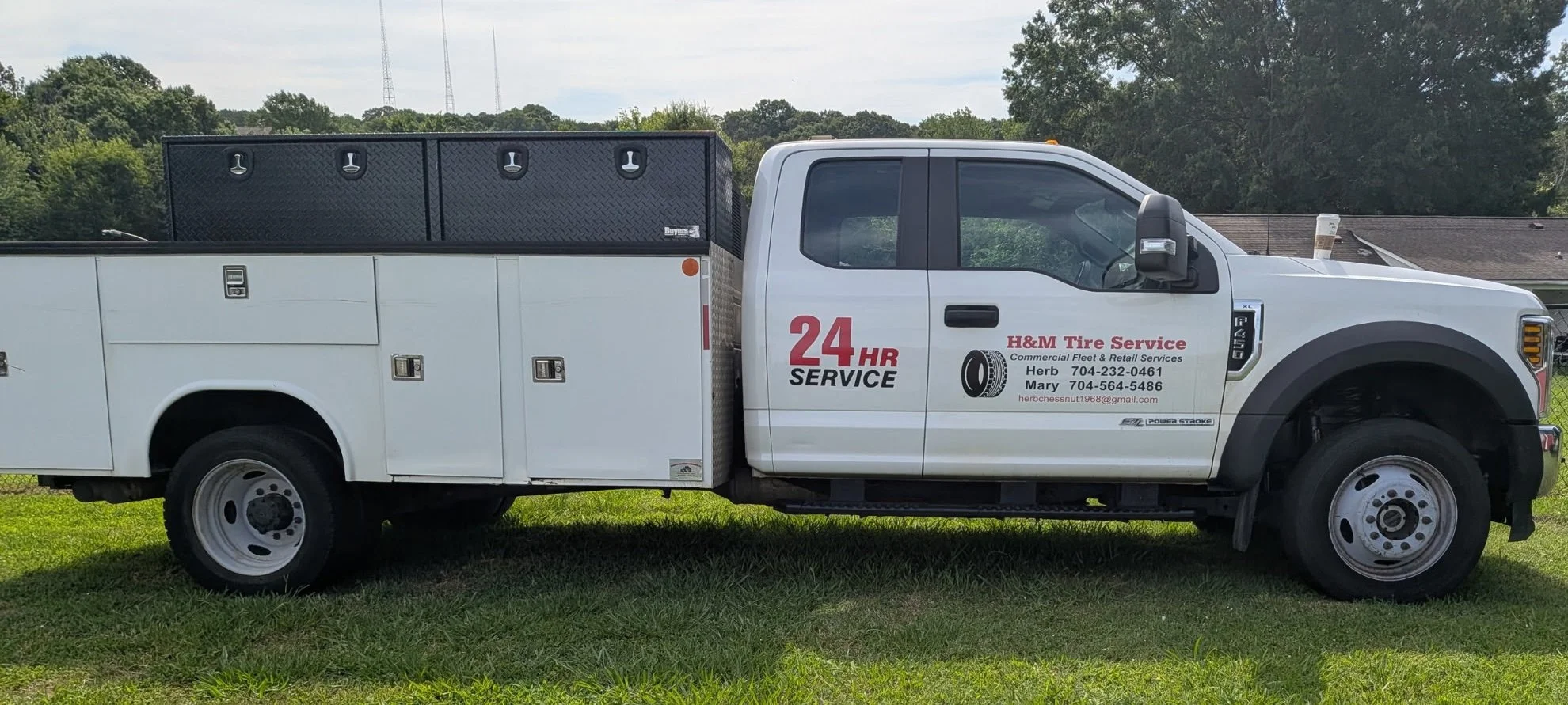 White service truck with signage for H&M Tire Service parked on grass, with a house and trees in the background.