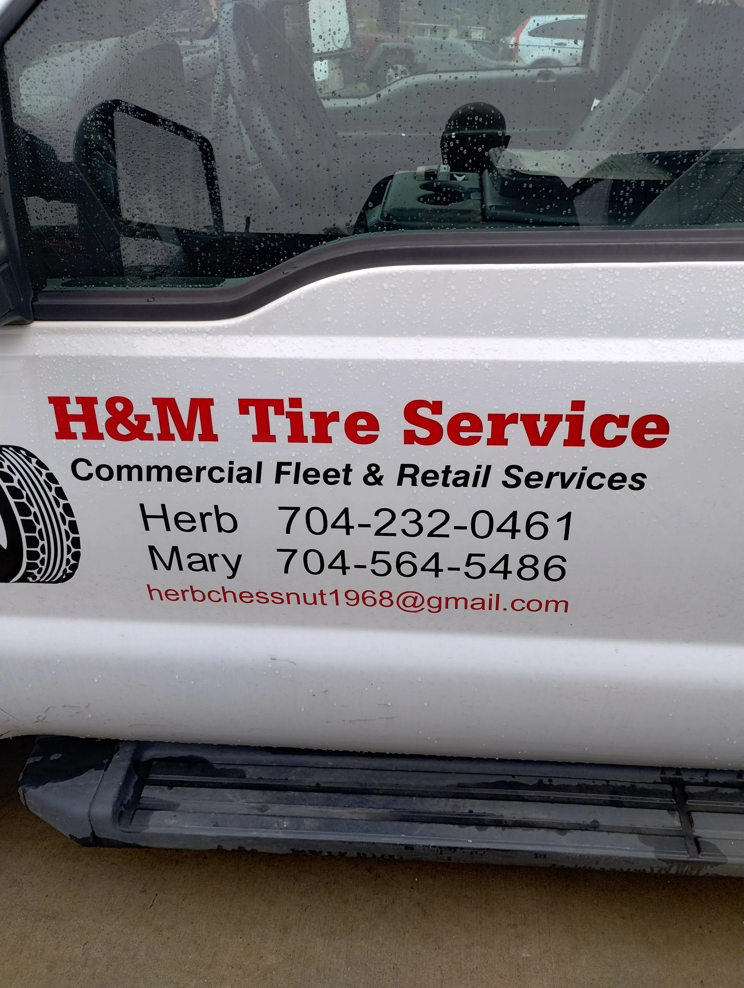A white vehicle with a sign for H&M Tire Service, showing contact information for Herb and Mary, on a concrete surface on a rainy day.