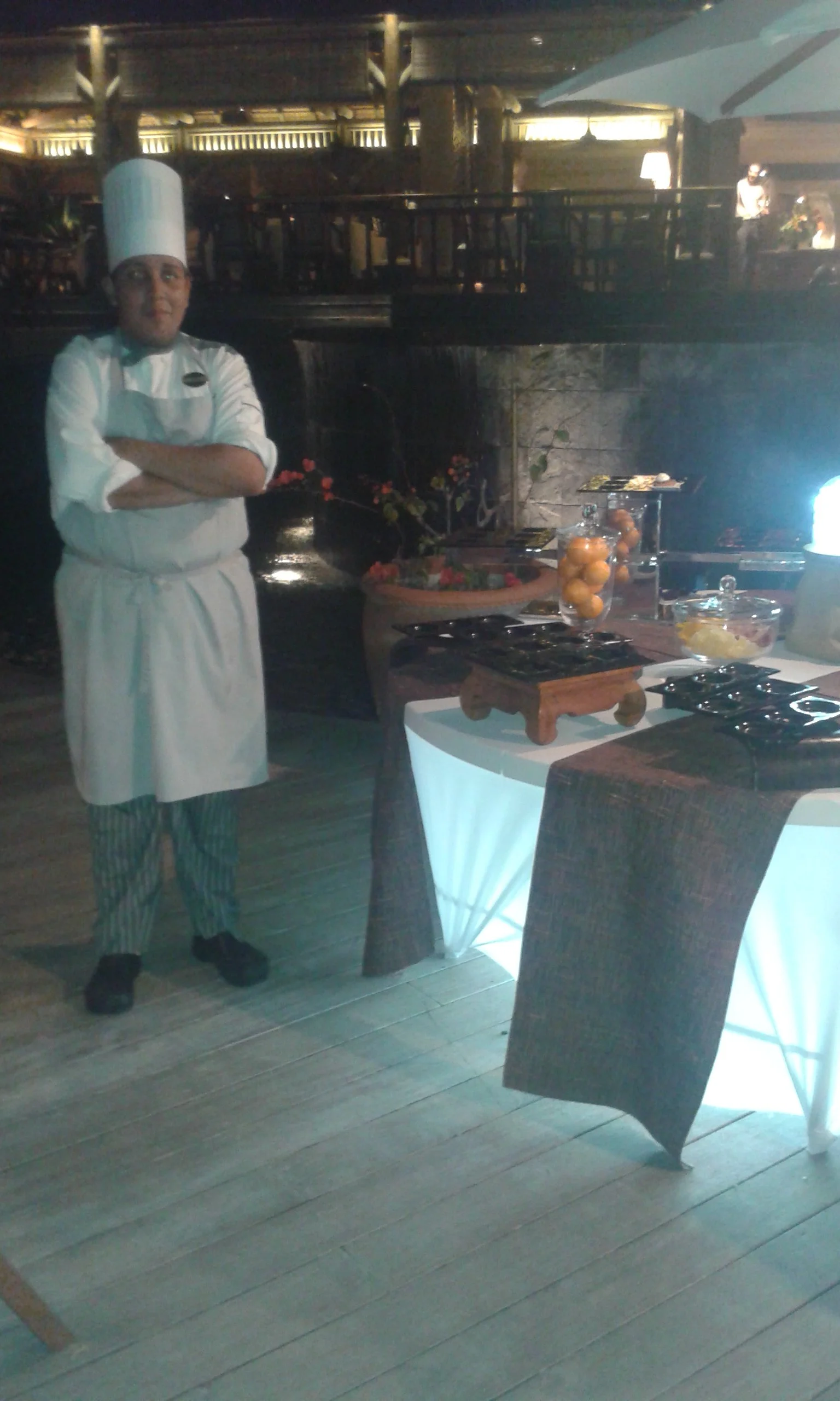 Chef in white uniform and tall hat standing beside a buffet table in a dimly lit restaurant