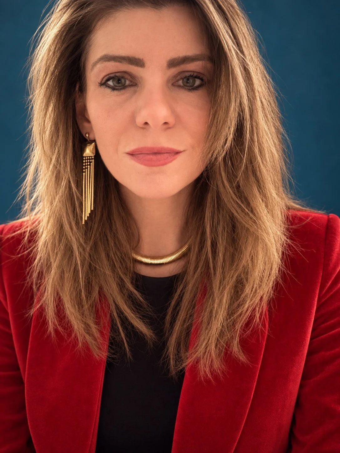 A woman with wavy, shoulder-length hair, wearing a red blazer, gold jewelry, and a black top, looking at the camera with a slight smile, set against a blue background.