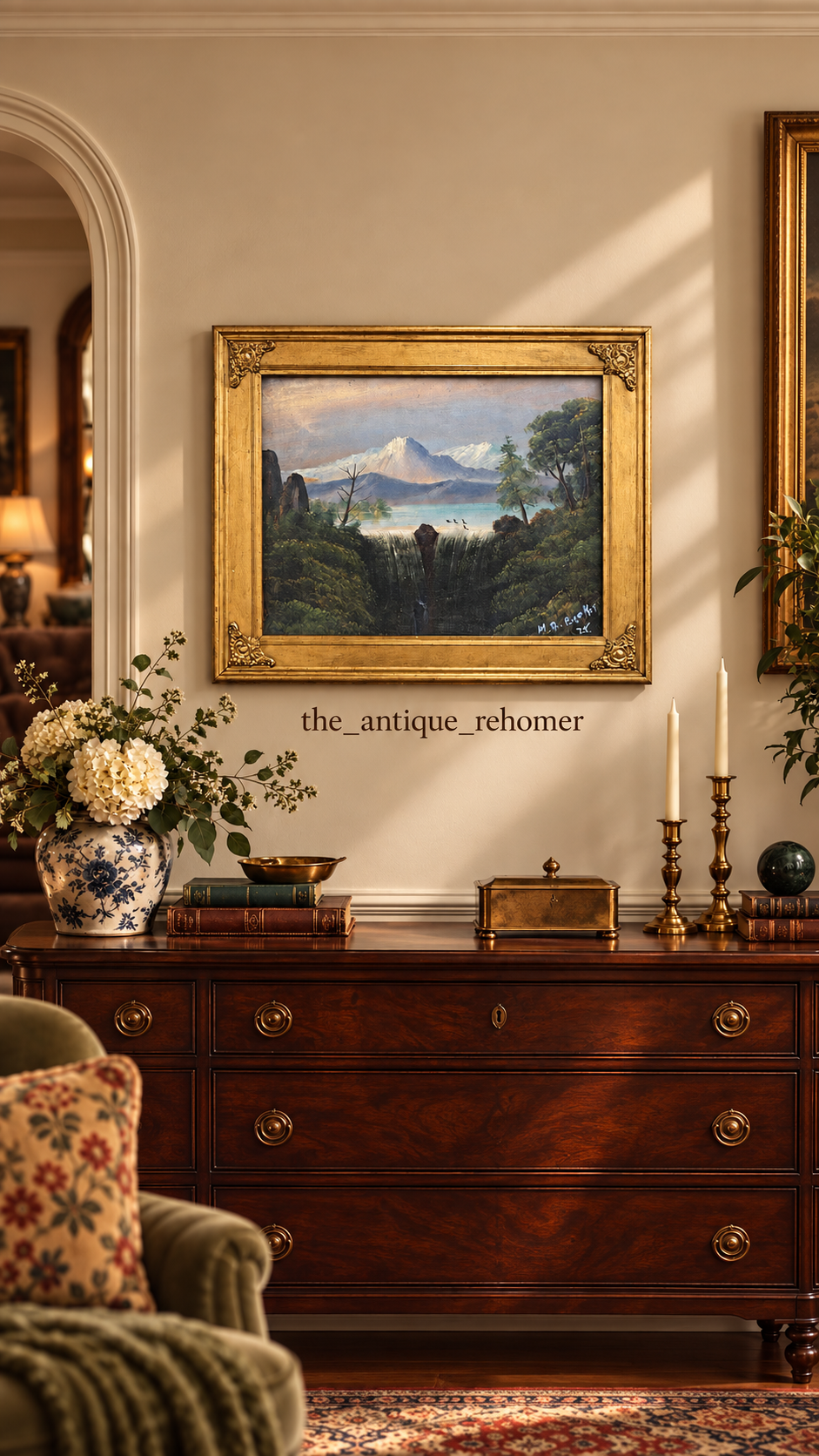 A classic wooden sideboard with a floral bouquet in a blue and white vase, green and brown books, a brass bowl, a gold box, and two gold candlesticks with white candles. A framed landscape painting of mountains, water, and trees hangs on the beige wa