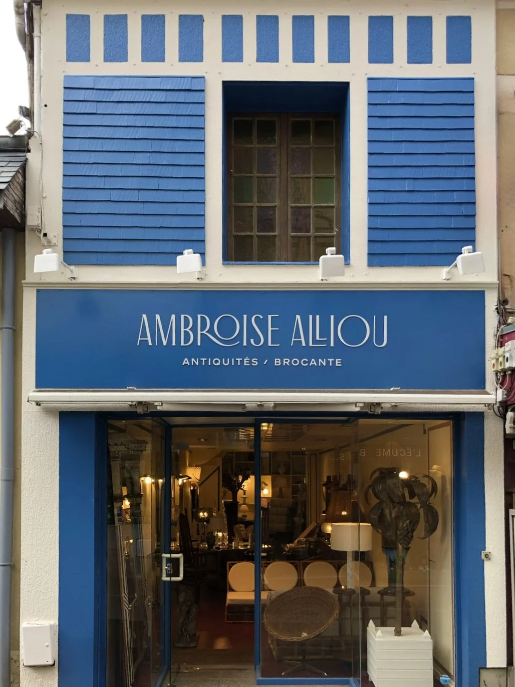 Façade d'une boutique avec un panneau bleu portant le nom 'Ambroise Aliou, Antiquités / Brocante'.