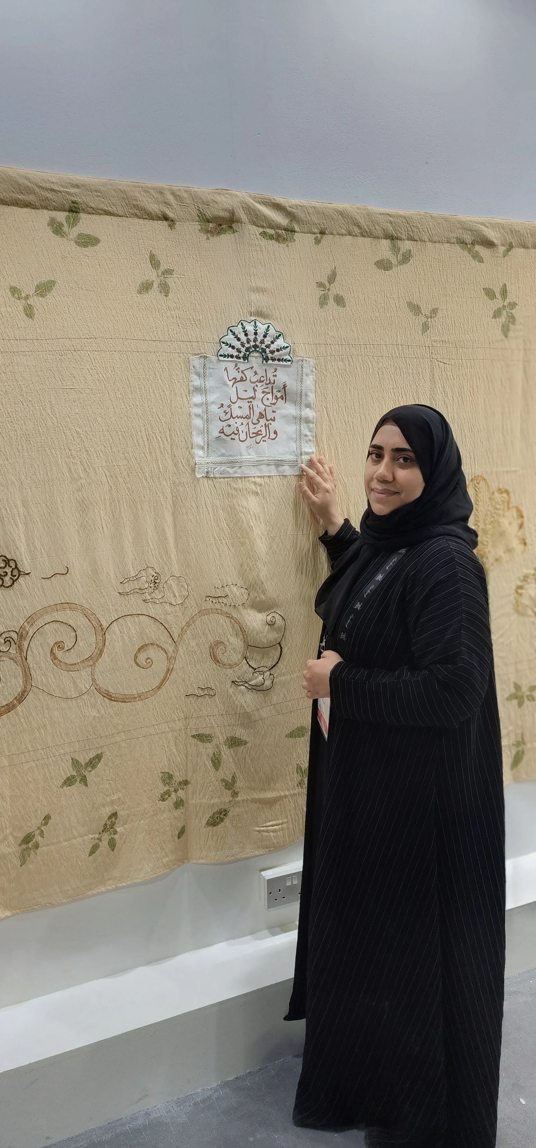 The artist standing next to a tapestry with Arabic calligraphy and decorative clouds. She is touching the tapestry and looking at the camera.