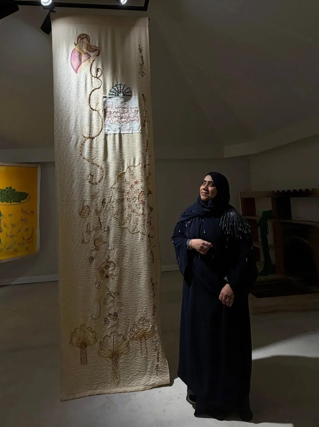 The artist stands in an art gallery, looking at a tall, textured fabric artwork with embroidery and drawing of a mermaid, trees, and Arabic script.