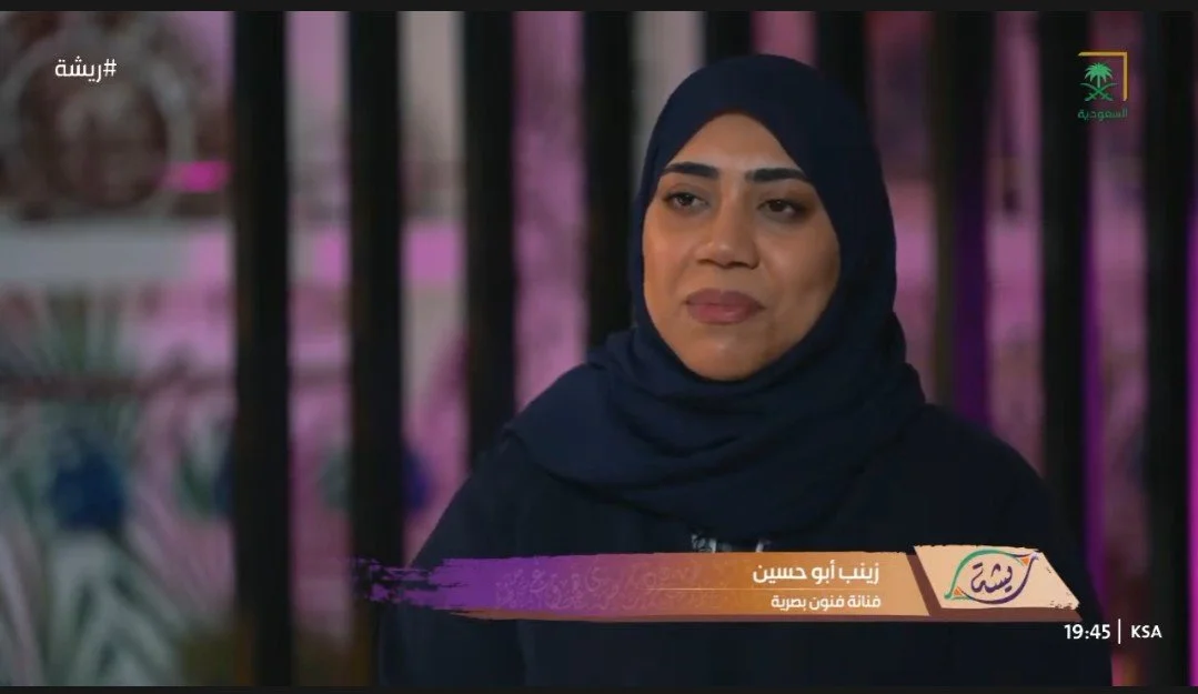 The artsit sitting in front of a blurred background with pink and purple hues, during a televised interview or segment in Saudi Arabia.