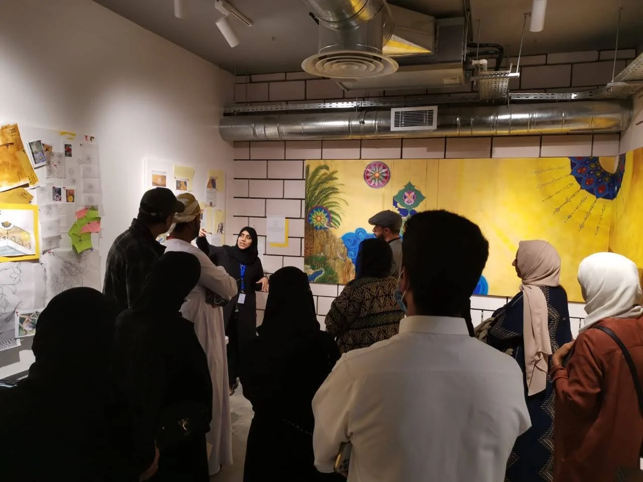 Group of people indoors, standing in front of colorful artwork on the wall, listening to an artist speaking during a guided tour or presentation.
