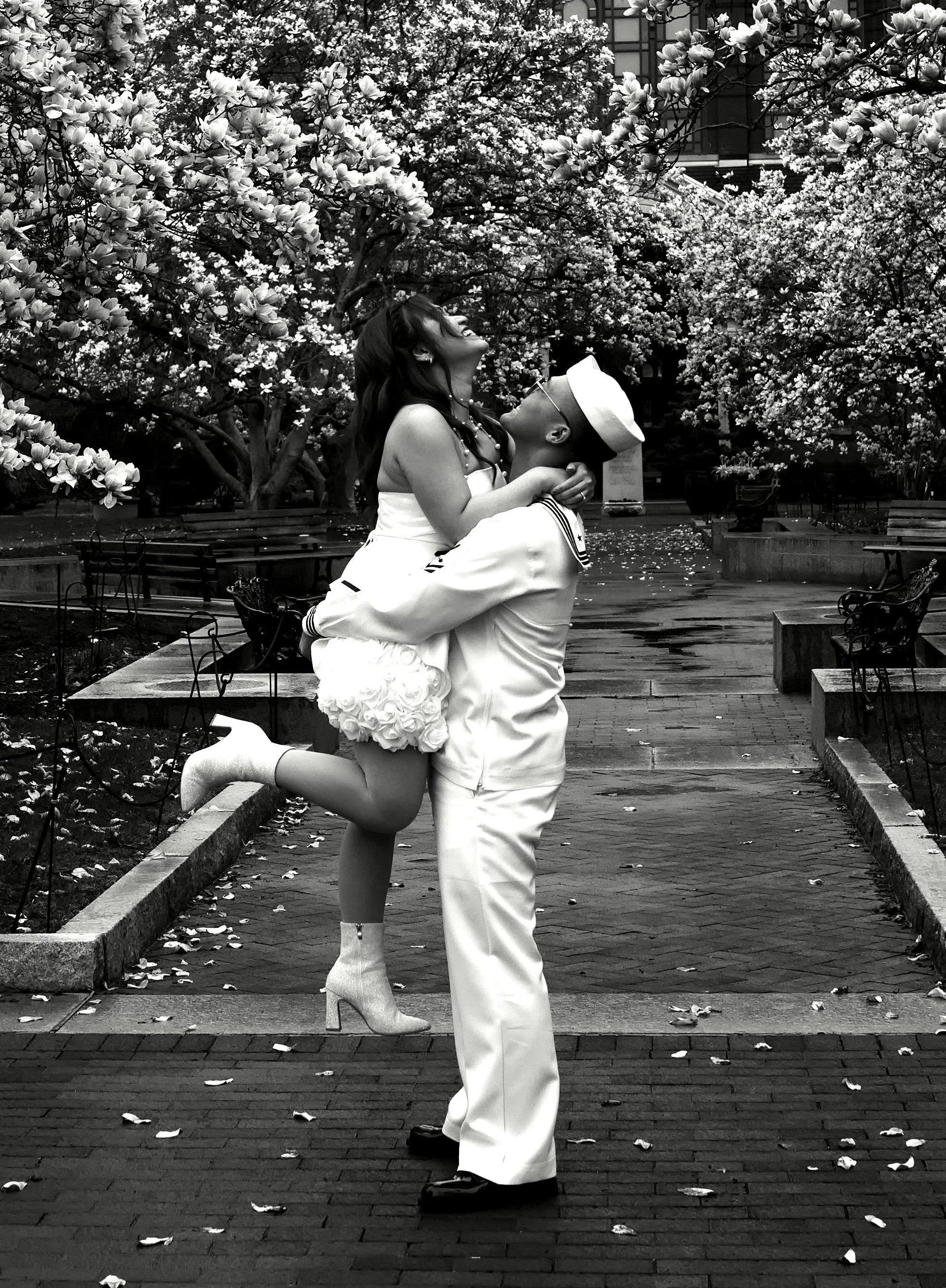 A person in a white outfit, including a sailor hat and jacket, is holding another person who is dressed in a floral dress and high-heeled boots, in a park with blooming trees, benches, and fallen leaves, in black and white.