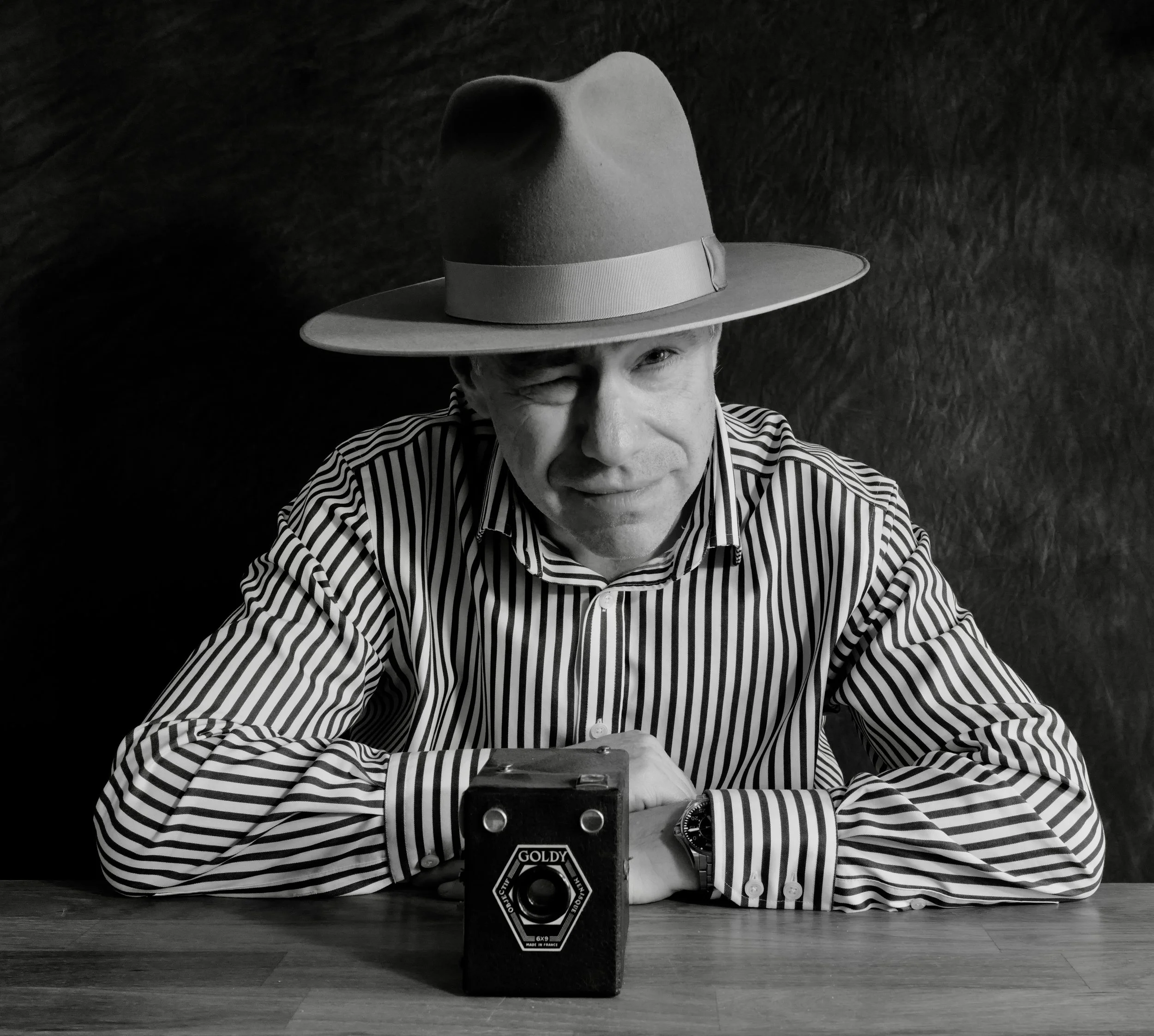 A person with a striped shirt, wearing a fedora hat, is sitting at a table, partially covering one eye with their hand. They are behind a vintage camera on the table, and the background is dark. The image is in black and white.