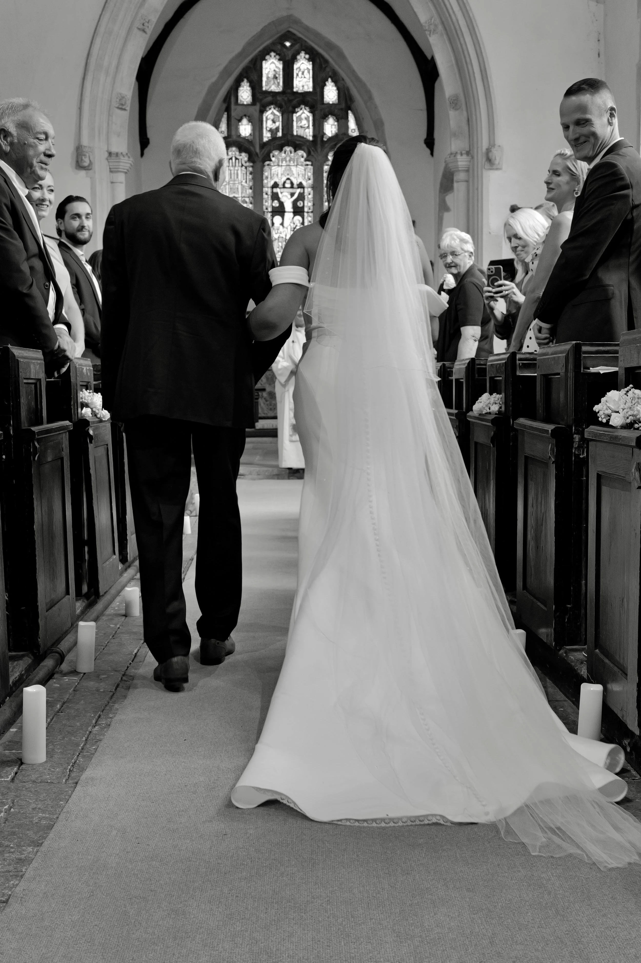 Black and white photo of a bride walking down the aisle of a church, accompanied by two men. Guests on either side of the aisle are smiling and taking photos. The church interior features stained glass windows.
