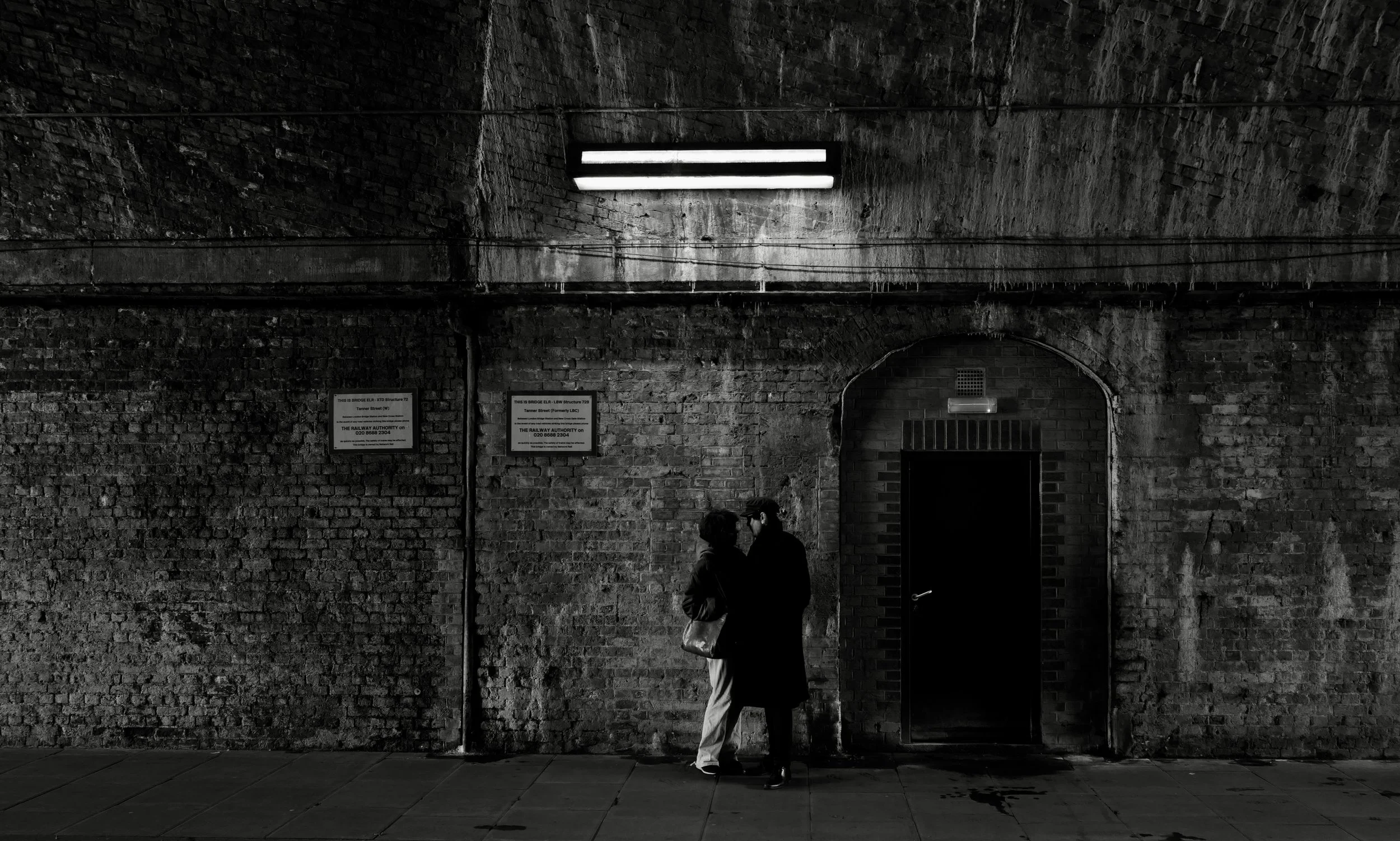 Two people standing close and talking against a brick wall with signs and a doorway, illuminated by a fluorescent light fixture in an underground or tunnel area.