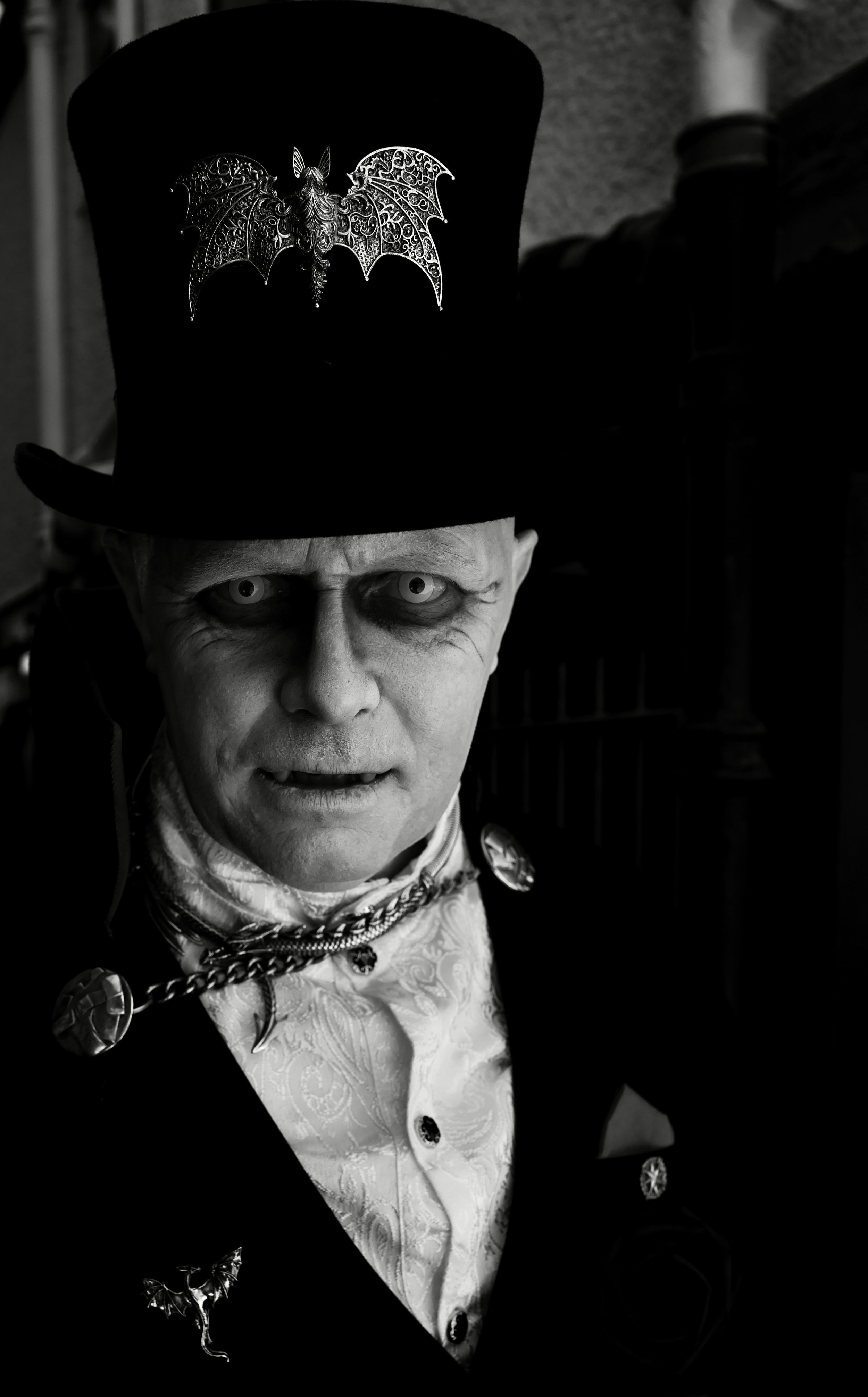 A person in gothic makeup wearing a top hat with a decorative emblem, a white shirt with intricate embroidery, and a chain with bat-shaped charms.