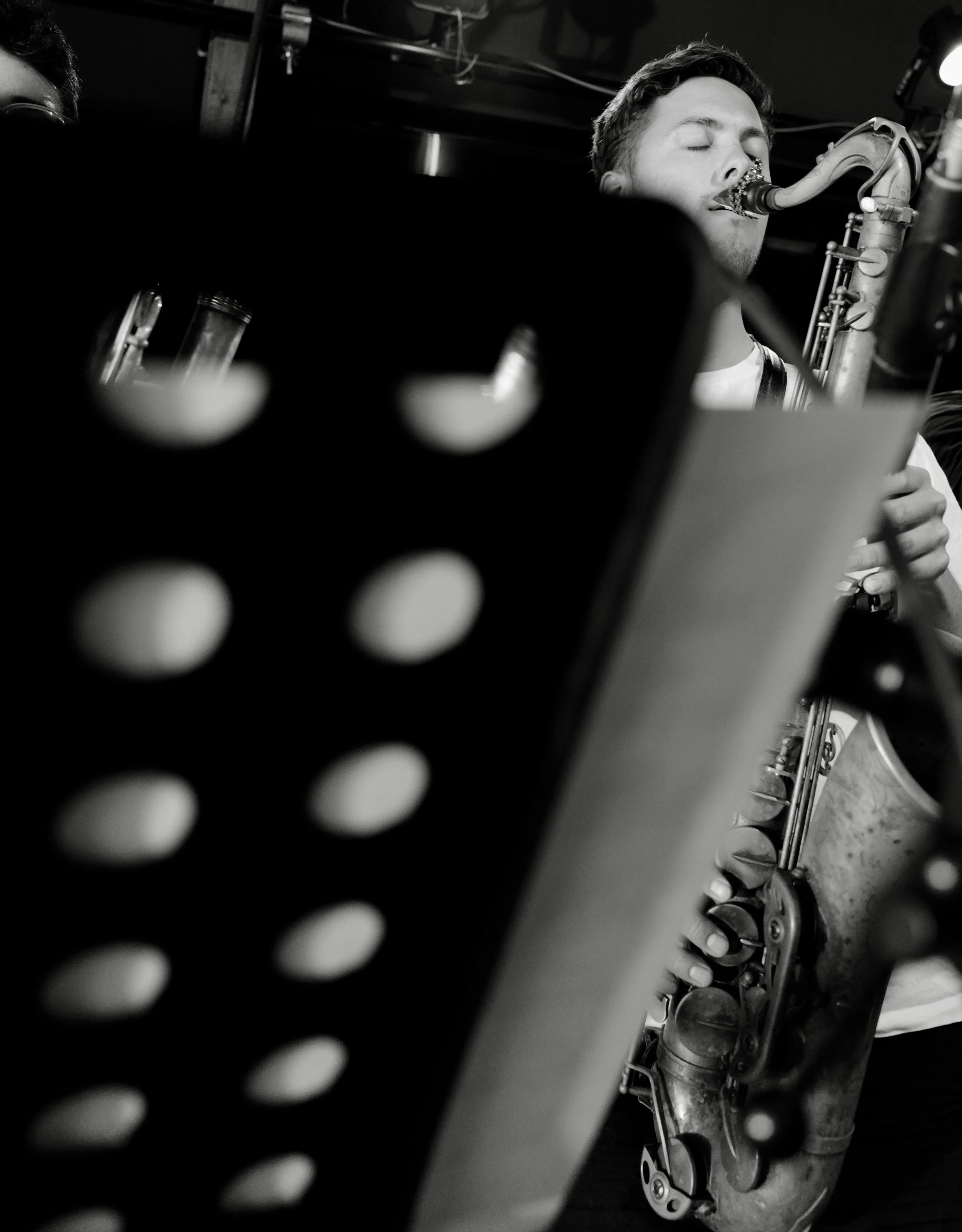 A black and white photograph of a young man with closed eyes playing a saxophone, viewed through a perforated surface that is out of focus in the foreground.