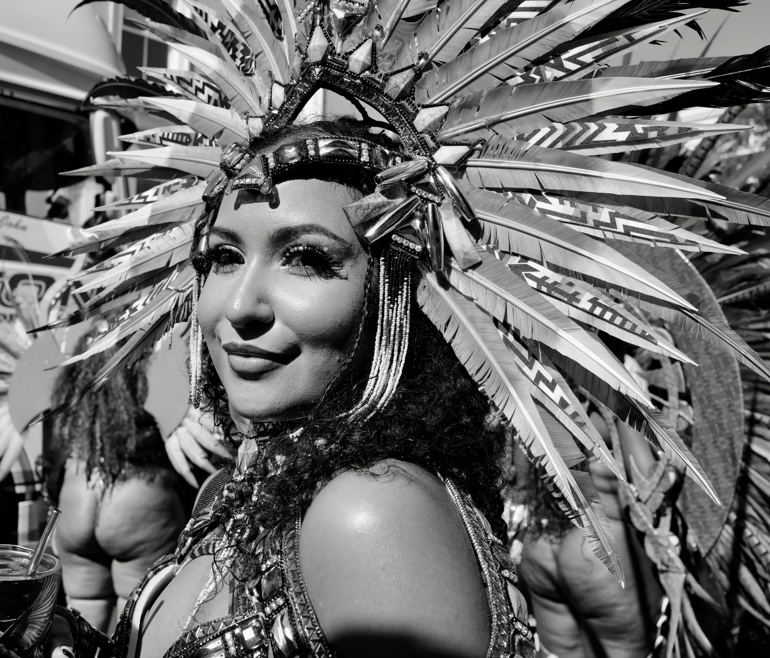 A woman dressed in a carnival costume with feathered headdress, sparkling embellishments, and intricate jewelry, looking to the side with a confident expression.