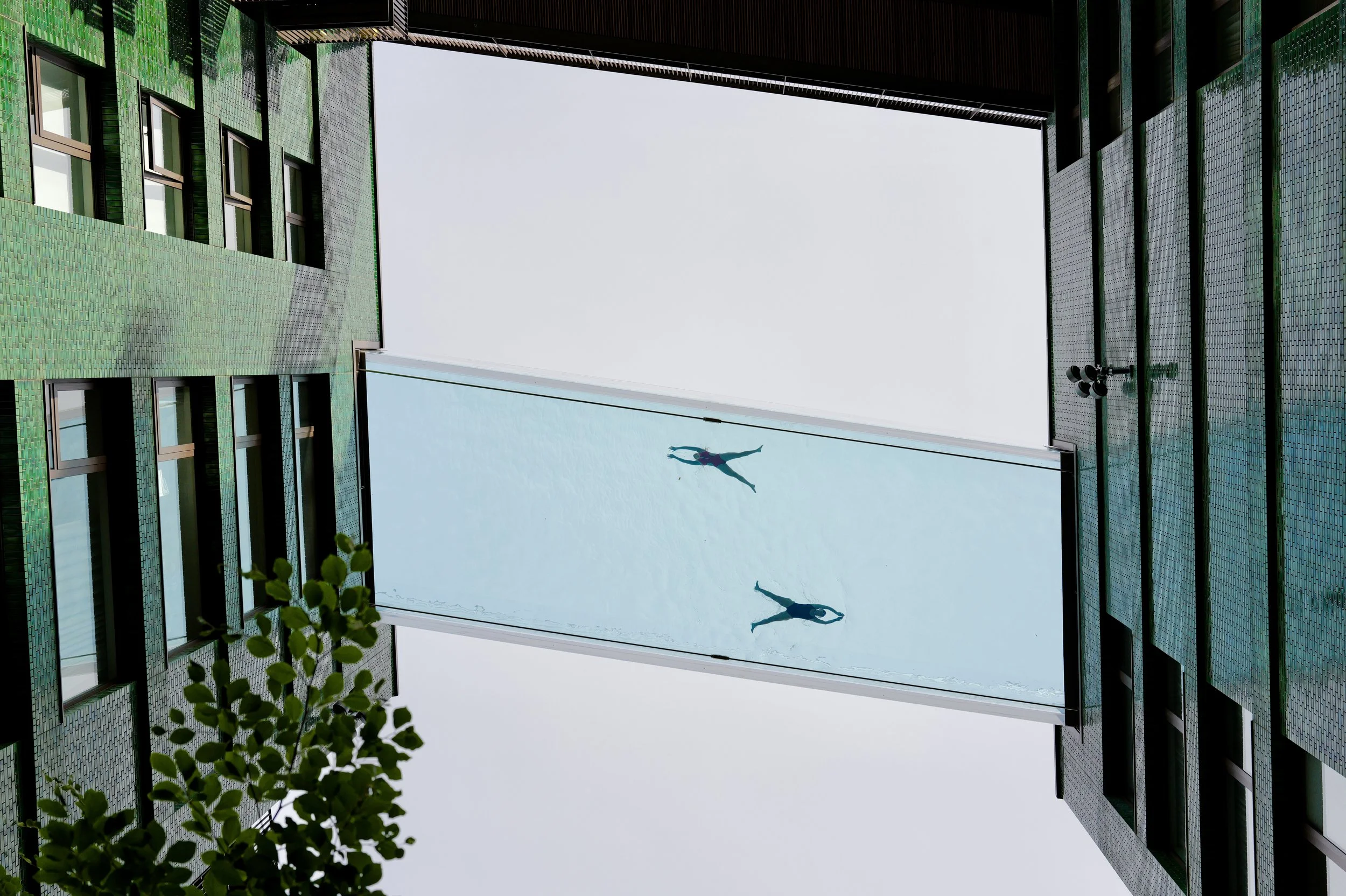 View of a modern building with green-tiled walls and large glass windows. A glass panel with reflections of two people in swimming suits hanging in the courtyard between the buildings. Some foliage is visible in the lower left corner.