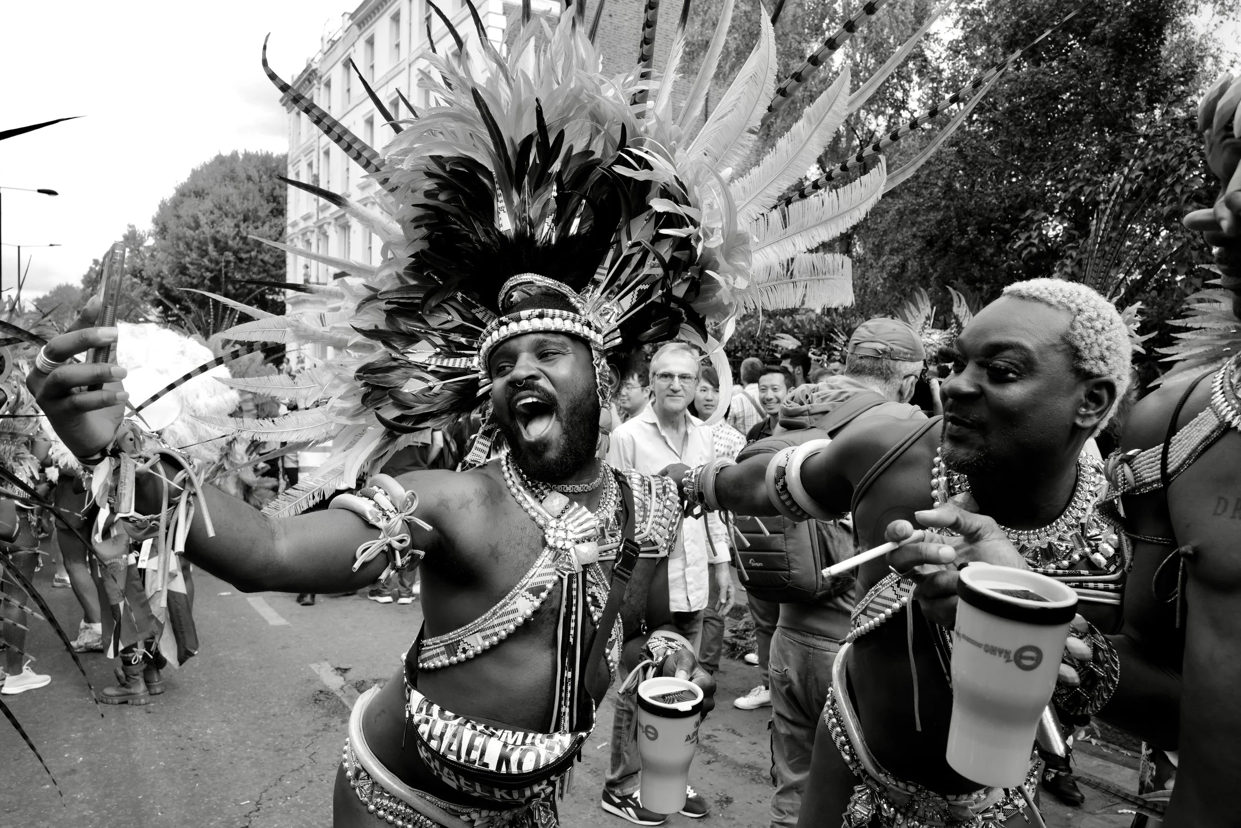 People at a parade, dressed in traditional African tribal attire, with one man wearing an elaborate feathered headdress, smiling and holding a parade cup. Others are dressed in beaded necklaces and accessories, surrounded by a crowd in an urban street setting.