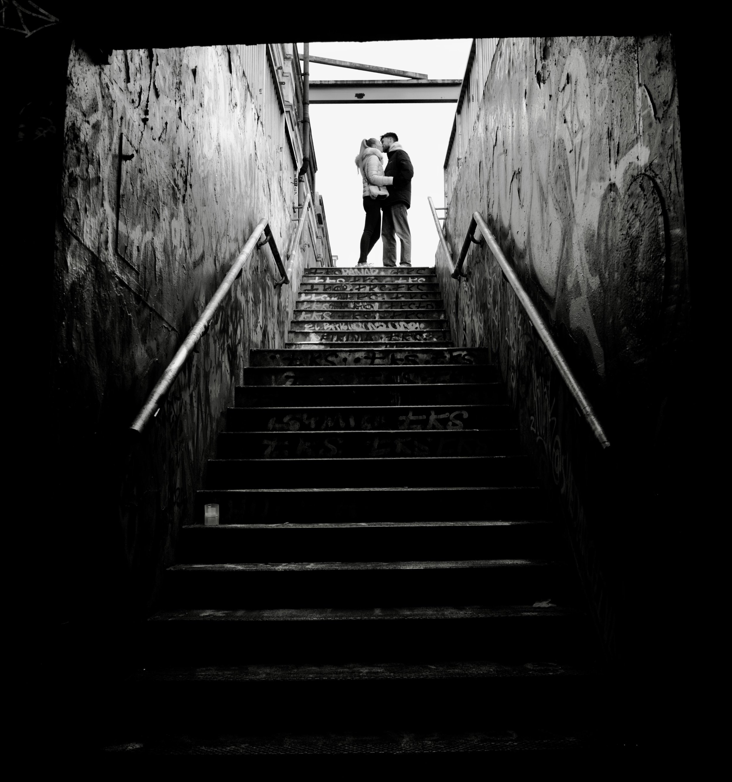 A black and white photo of a couple kissing on a staircase between two walls with graffiti. The couple is wearing winter jackets, and there is a light source behind them, creating a silhouette effect.