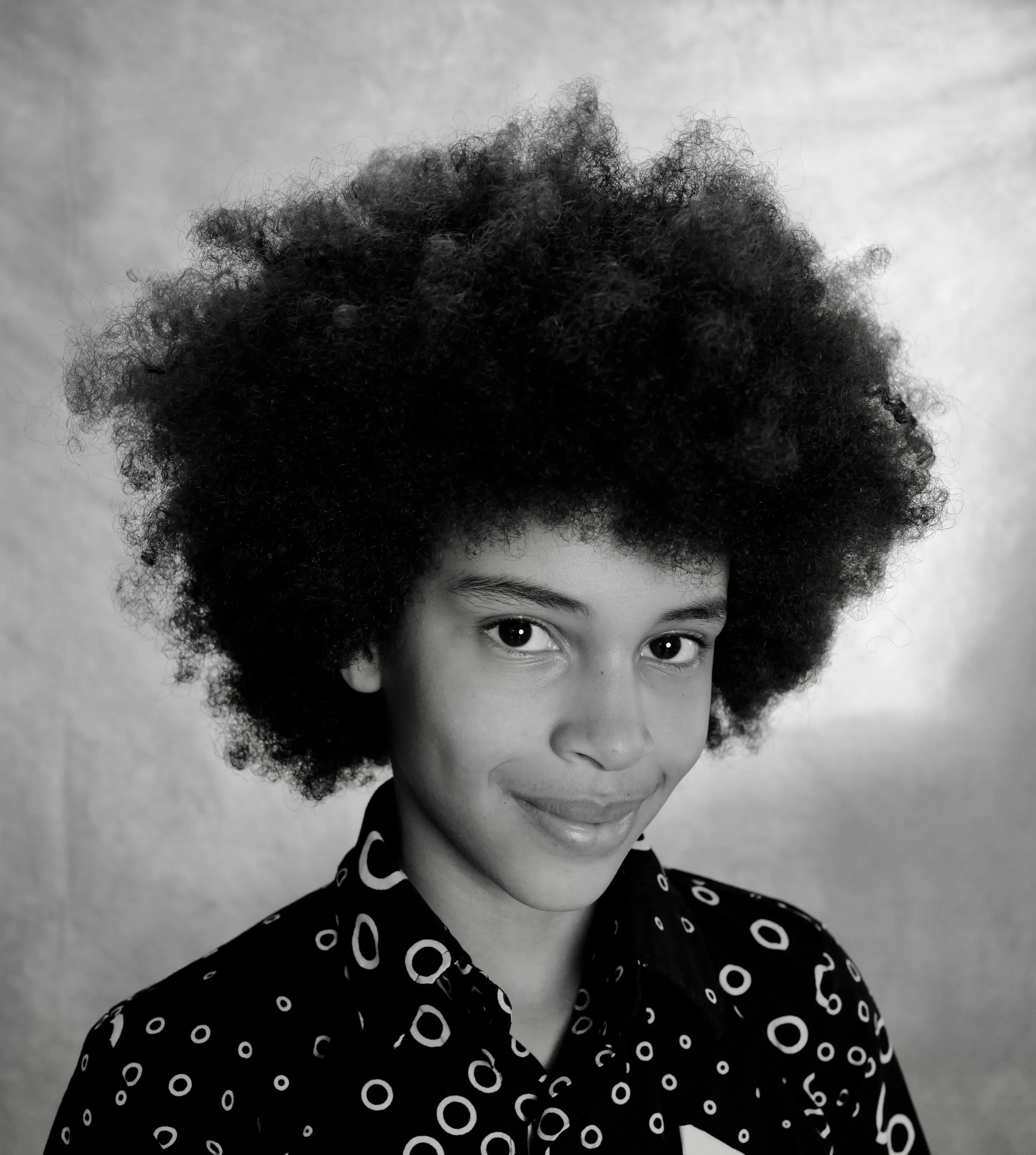A black and white photo of a woman with natural curly hair, slightly smiling, wearing a dark top with circular patterns.