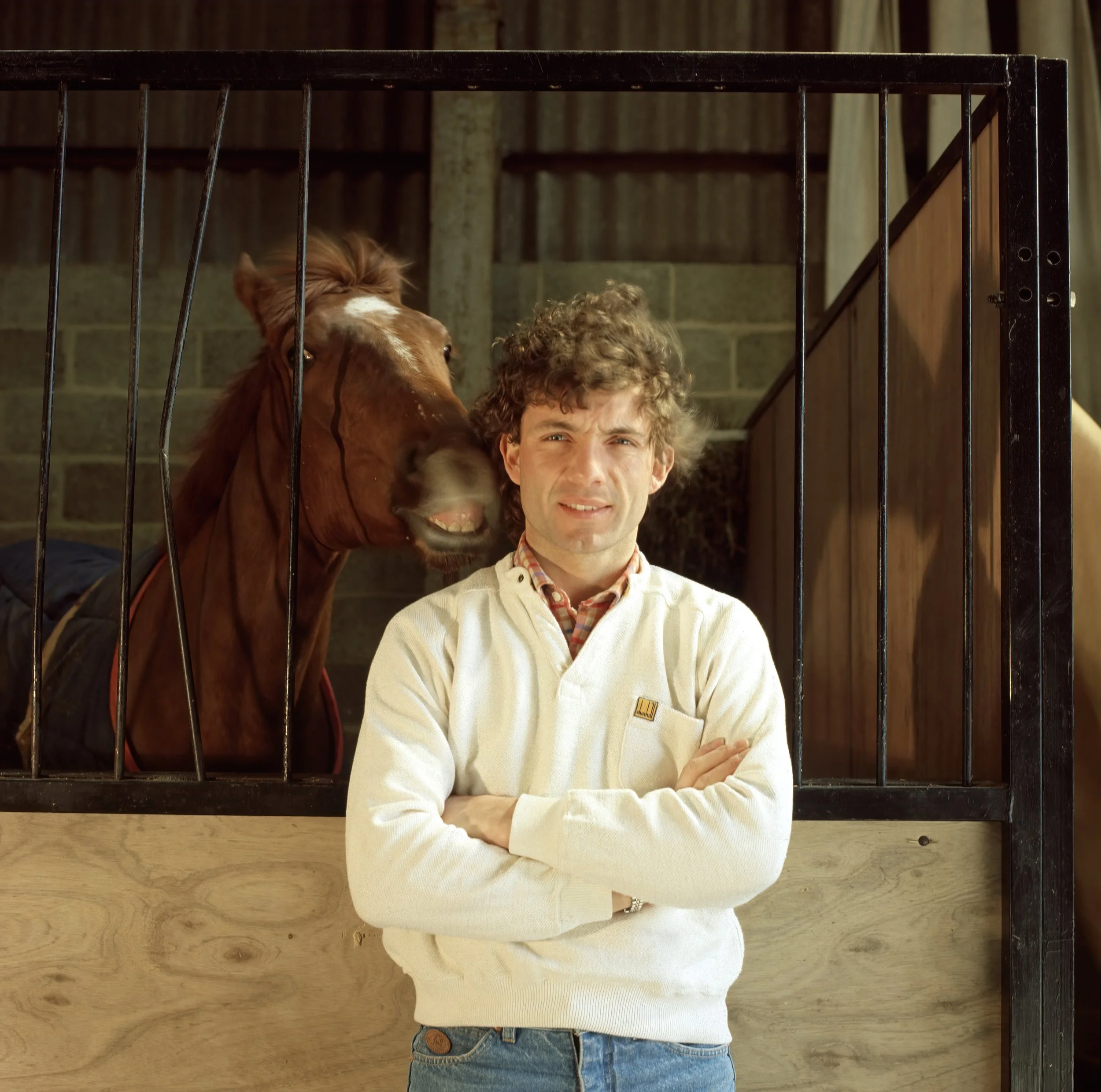 Partially visible person with arms crossed, wearing a white sweater and jeans, standing in front of a wooden and metal background, with a horse and a child behind bars.