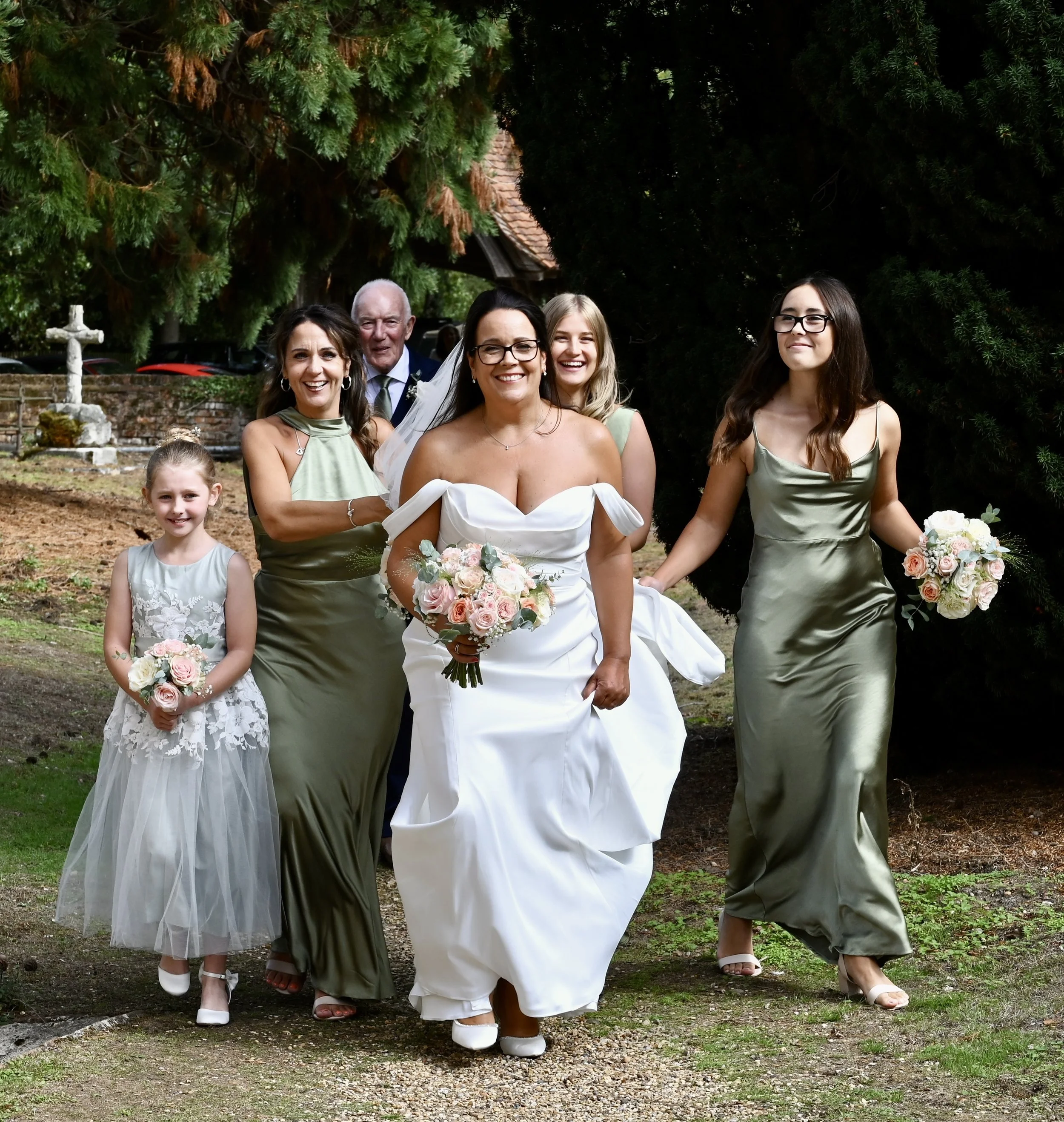 A group of people walking outdoors in a garden during a wedding or celebration. The group includes women, a man, and children, dressed in formal and semi-formal attire, with some holding bouquets of flowers. There are large green trees and a brick structure in the background.