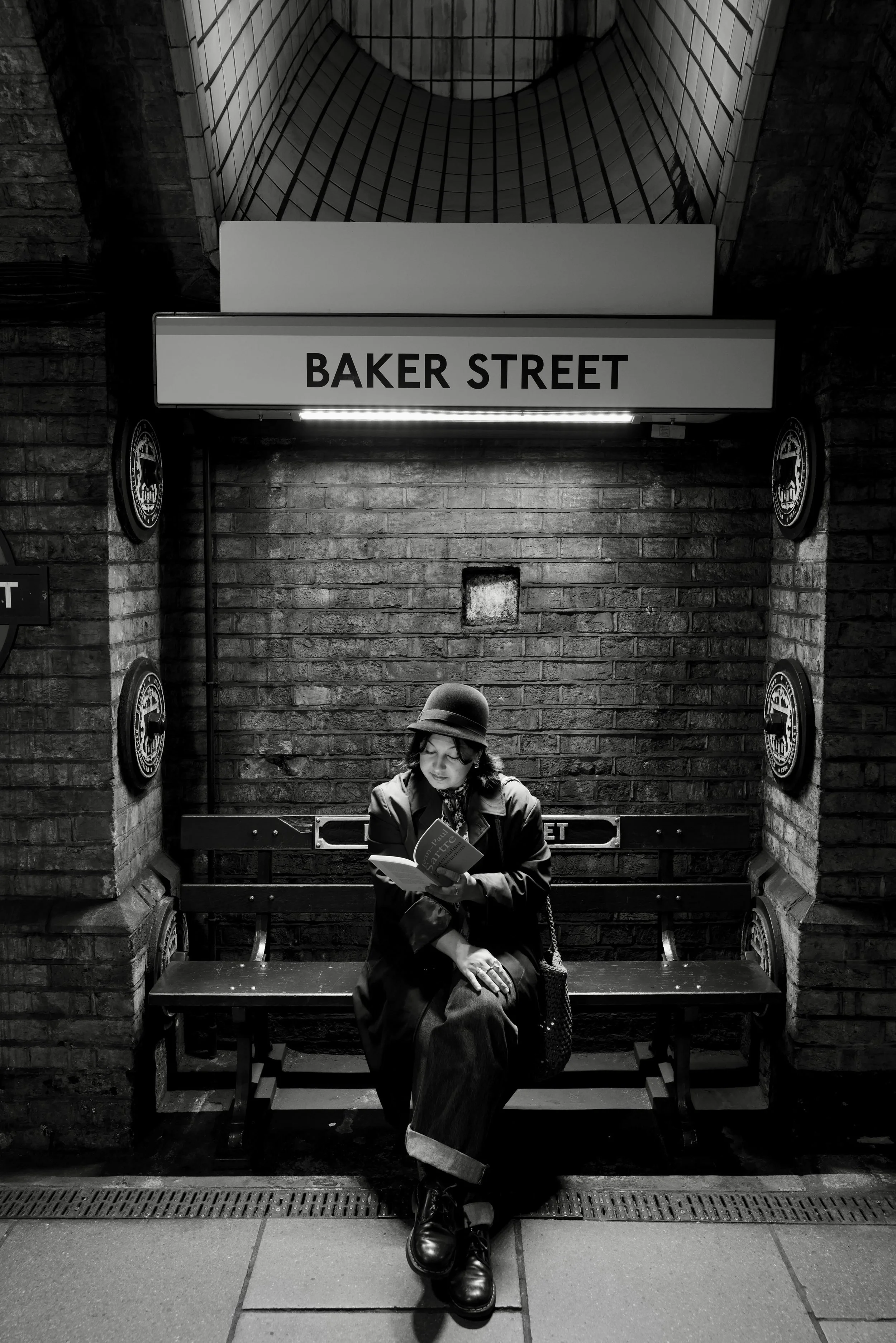 A woman in a coat and hat sitting on a bench reading a book in a subway station. There are signs and brick walls behind her, with a sign that reads 'Baker Street'.