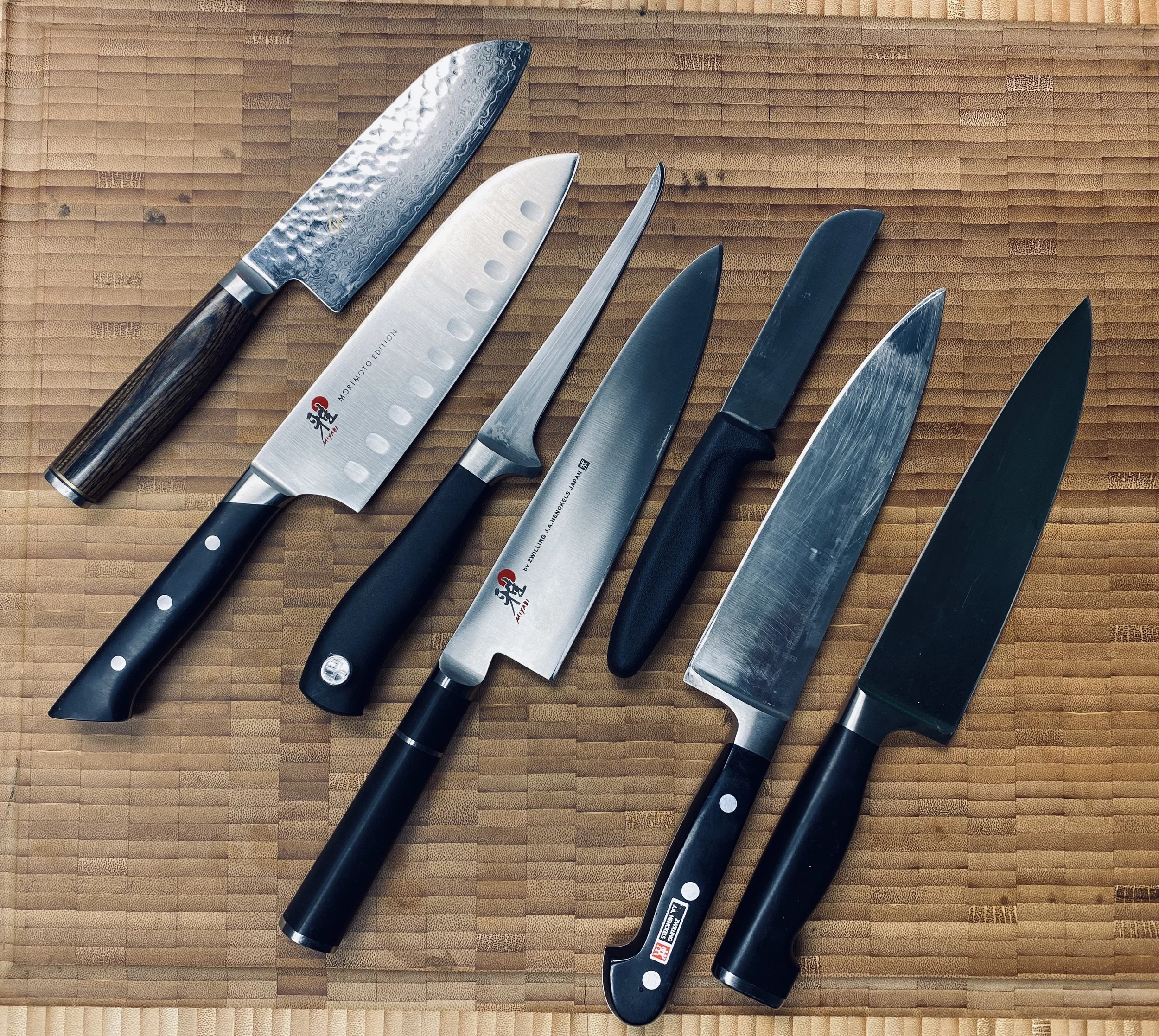 Set of six kitchen knives with black handles on a wooden cutting board.