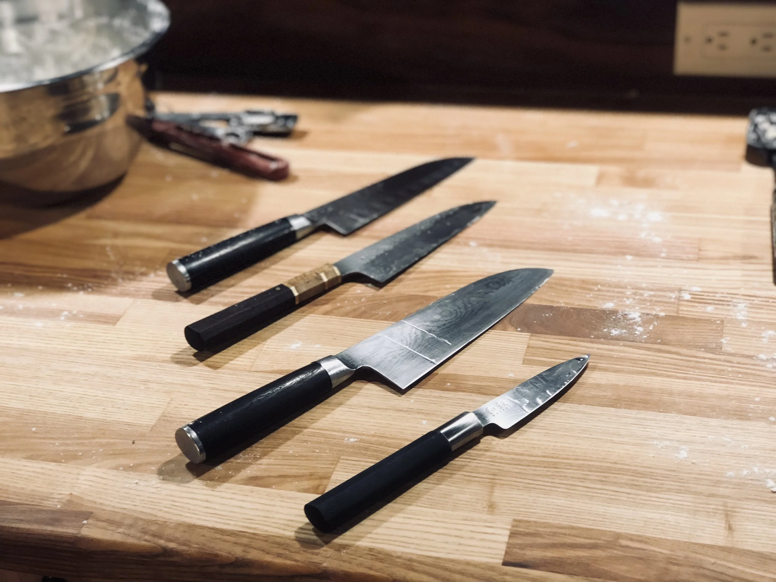 A wooden kitchen countertop with three knives laid out, a pot, a pair of scissors, and an electric outlet in the background.