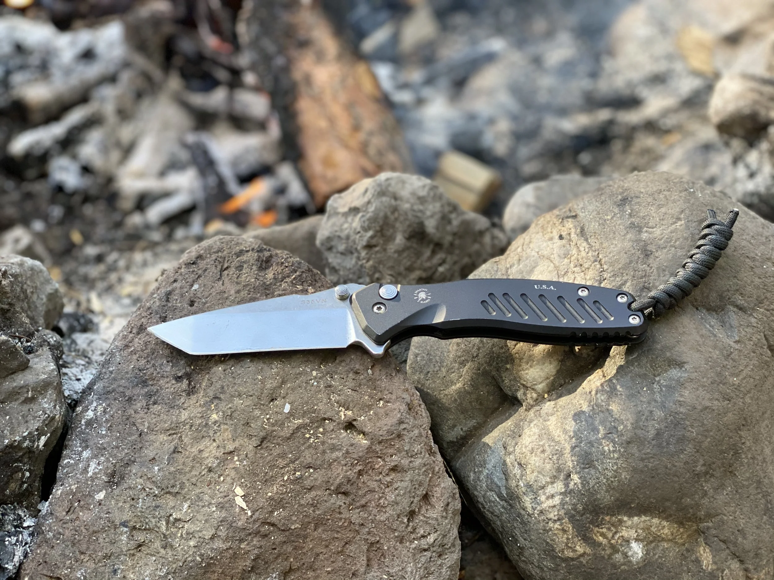 Close-up of a folding pocket knife with a black handle resting on rocks near a fire with gray stones and glowing embers.