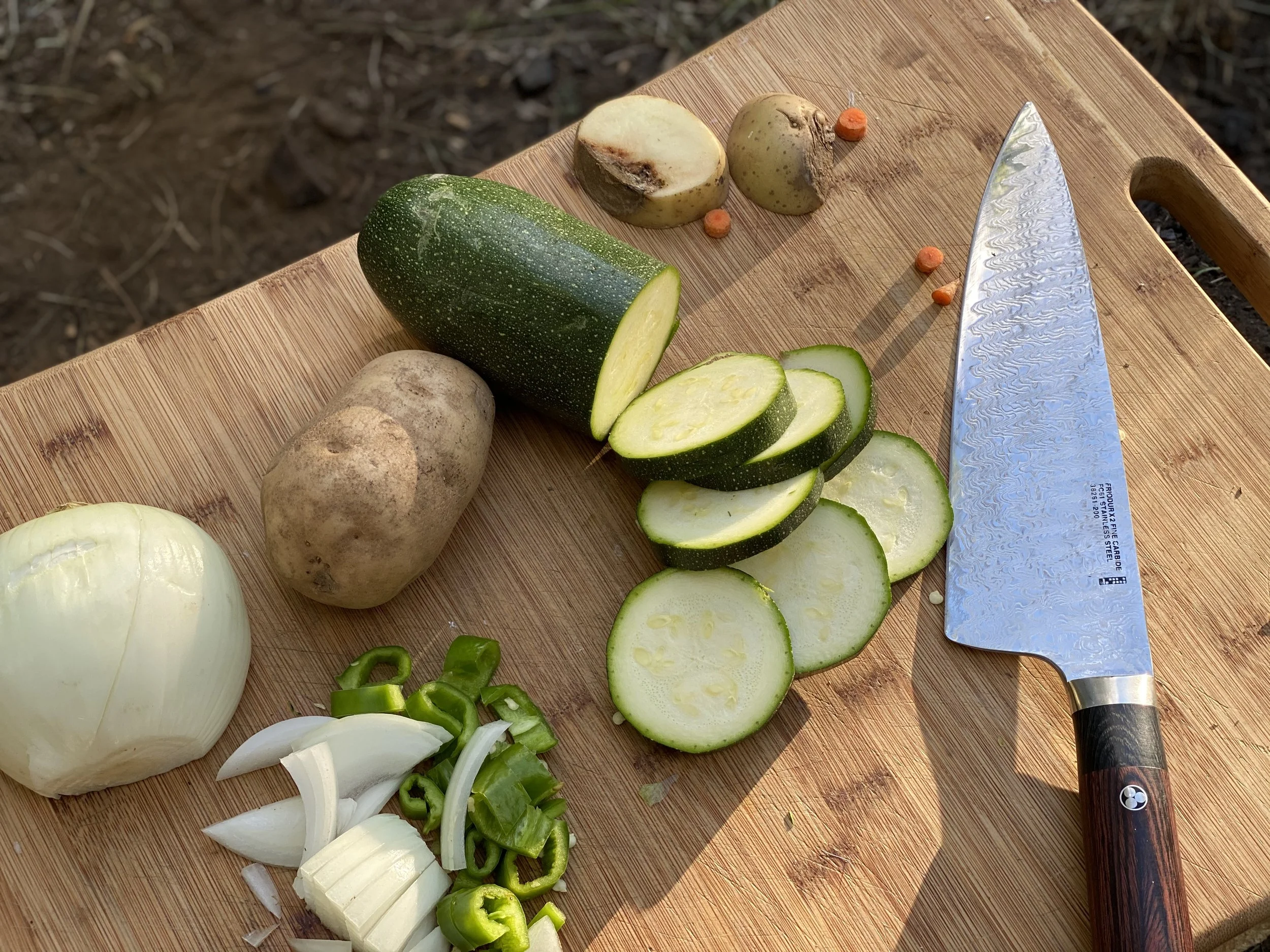 A bamboo cutting board with sliced zucchini, whole potatoes, chopped onions, and green bell pepper slices, with a knife.