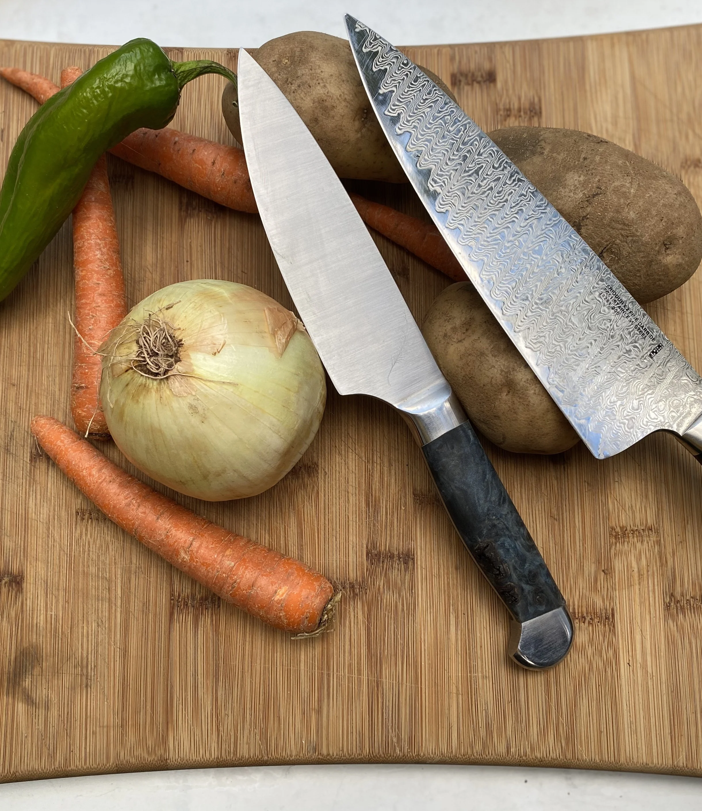 A cutting board with a green chili pepper, carrots, an onion, and two potatoes, along with a chef's knife with a Damascus pattern on its blade.