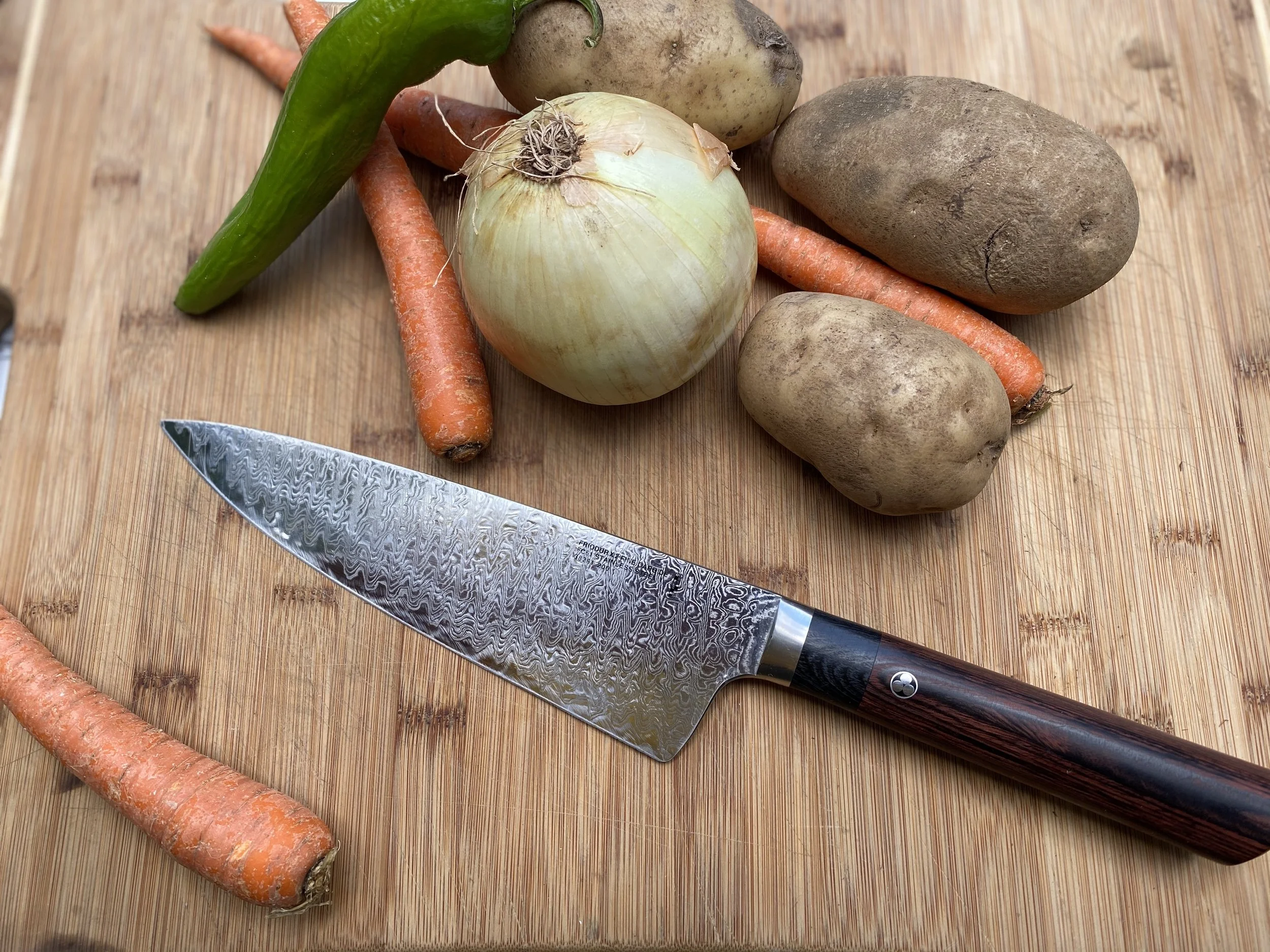 Fresh carrots, an onion, potatoes, a zucchini, and a knife on a wooden cutting board.