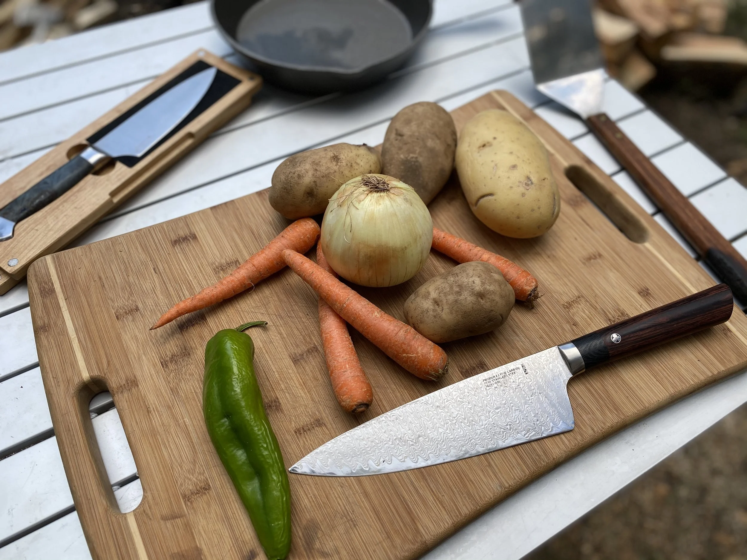 Fresh vegetables, including potatoes, carrots, an onion, and a green chili pepper, are arranged on a wooden cutting board with a knife and a black pan in the background.