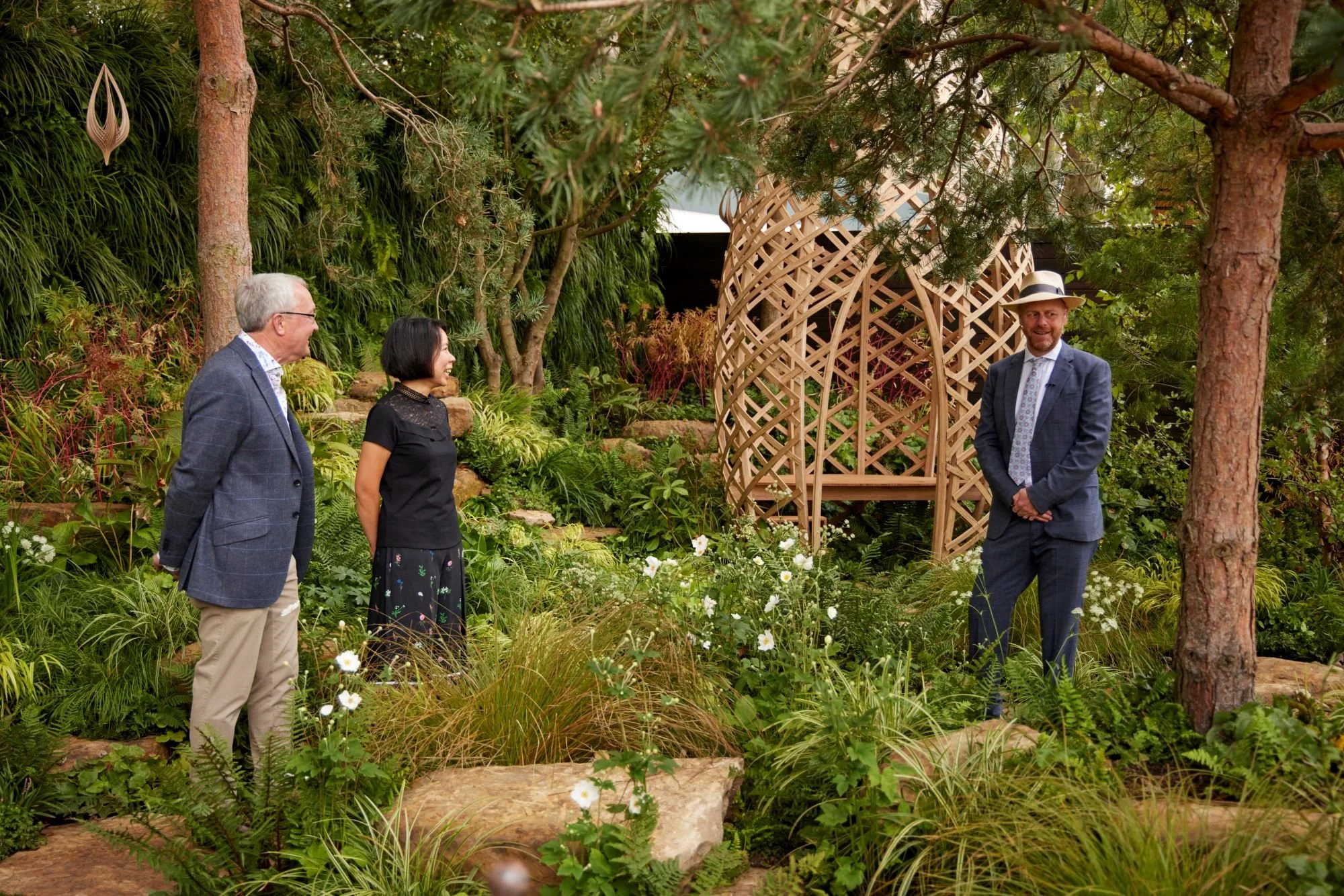 Four people are standing in a lush garden with a decorative wooden structure in the background. They are all dressed in business casual attire and are smiling, engaged in conversation. There are trees, green foliage, and flowering plants surrounding them.