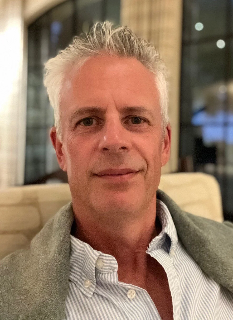 A middle-aged man with gray hair and fair skin smiling, sitting indoors near a window.
