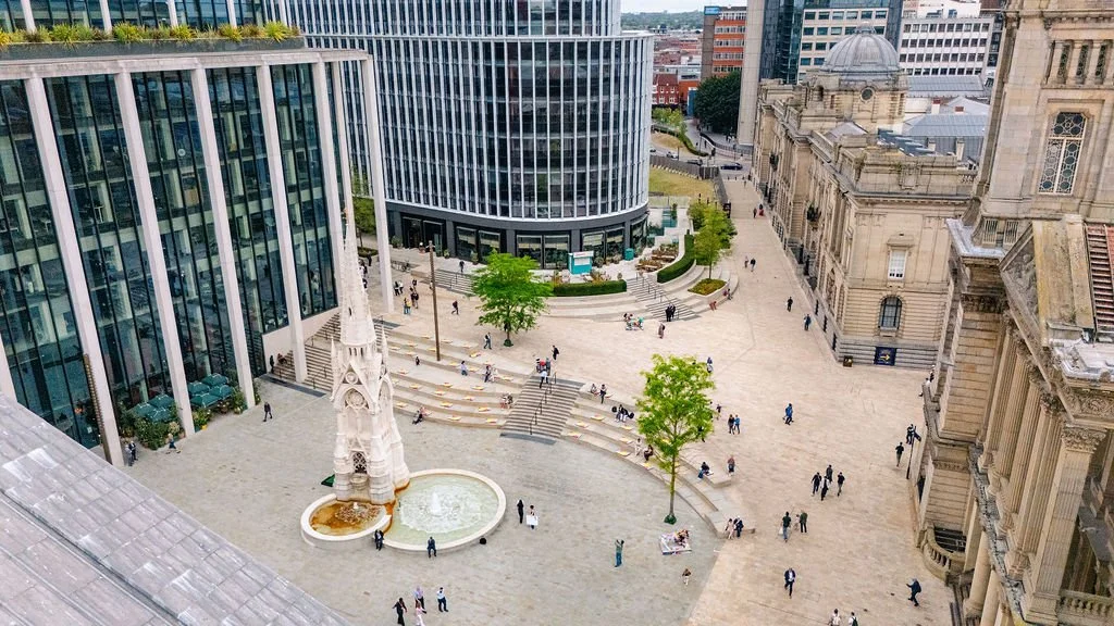 View of a city square with modern and historic buildings, a white church steeple, and people walking and sitting around the area.