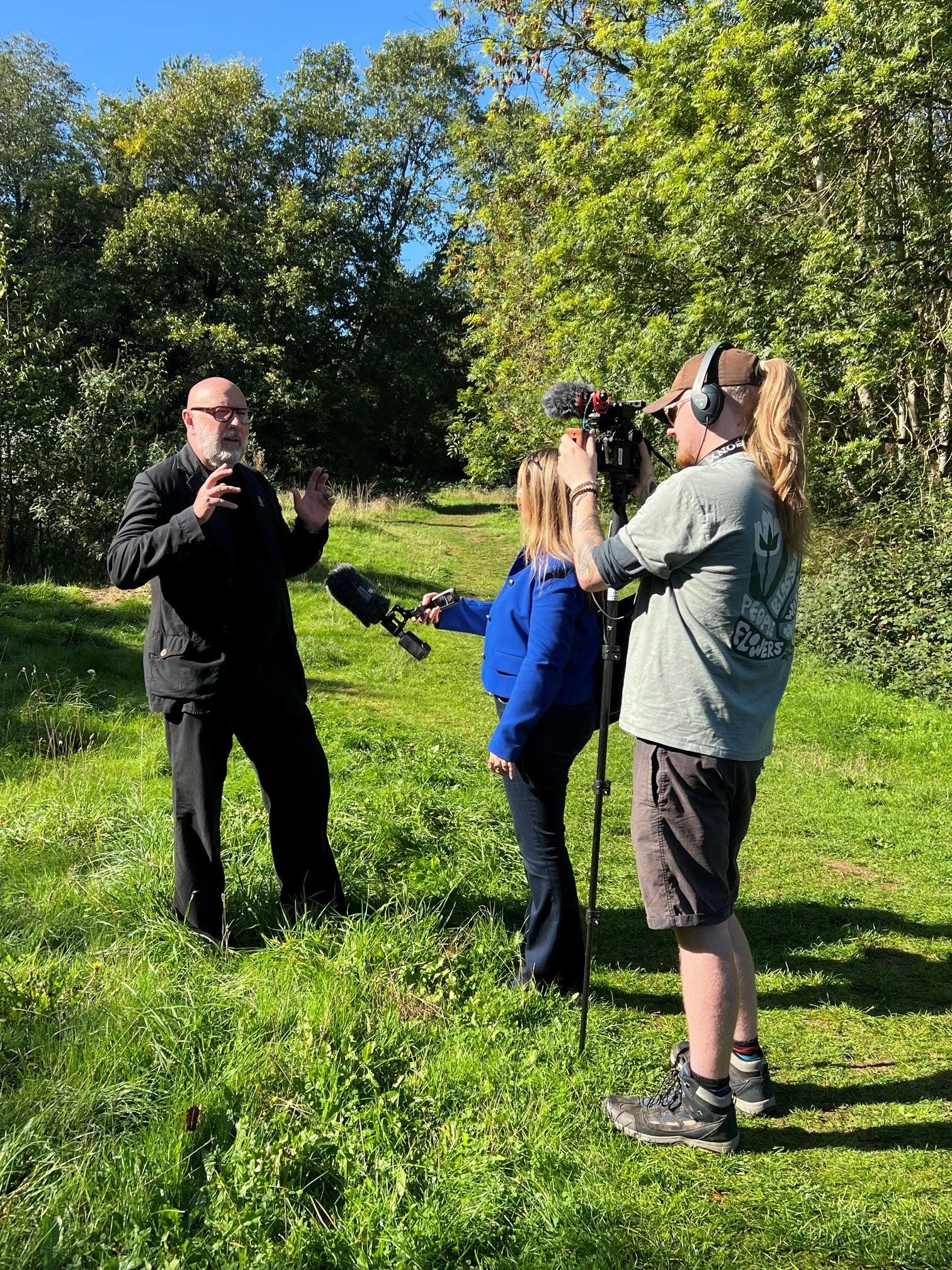 A man with glasses and a beard wearing black clothing is speaking to a camera crew in a grassy outdoor area with trees. The crew includes a woman with blonde hair in a blue jacket and a man with long hair, a cap, and headphones operating the camera. The scene is sunny with a clear blue sky.