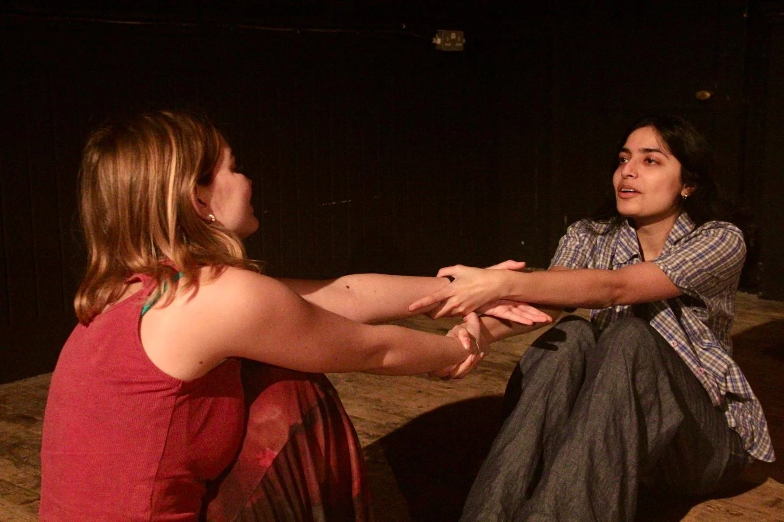 Two women are sitting on the floor, holding hands and engaging in a conversation or emotional exchange in a dimly lit room with dark walls.