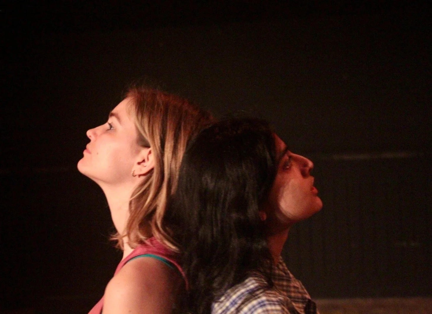 Two women standing back to back with their heads resting against each other in a dark setting.