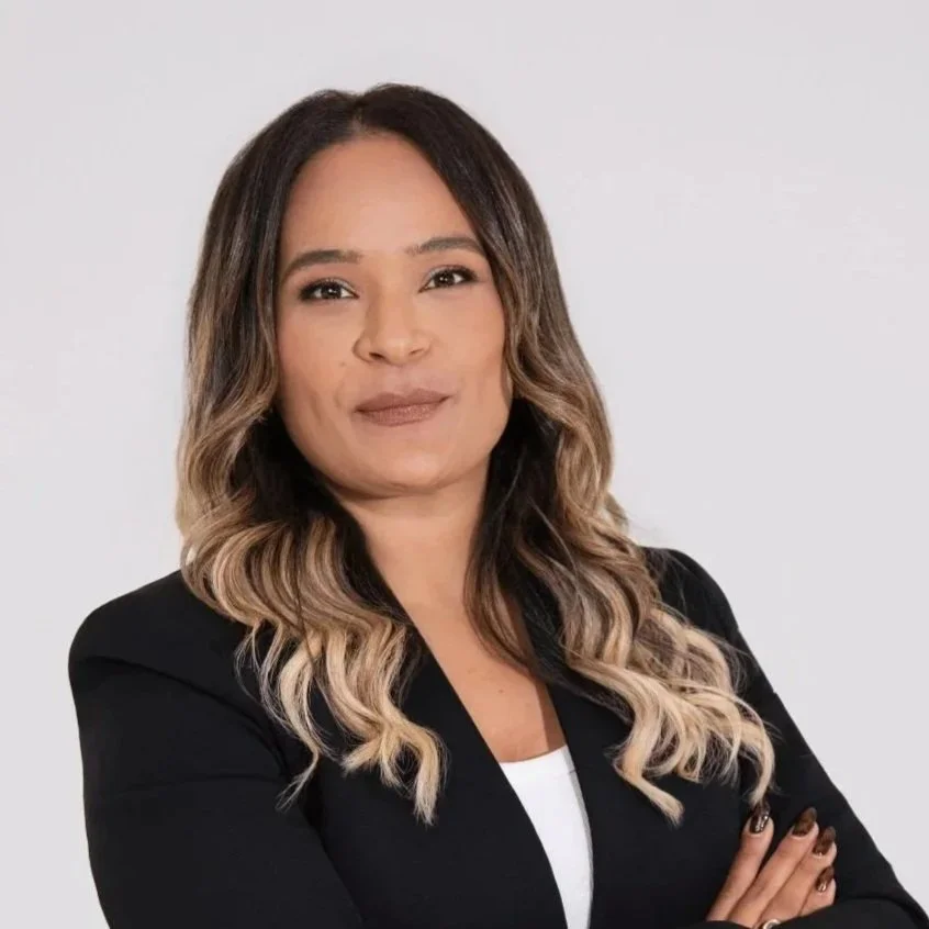 Professional woman with long highlighted hair wearing a black blazer and white top, posing with folded arms against a light gray background