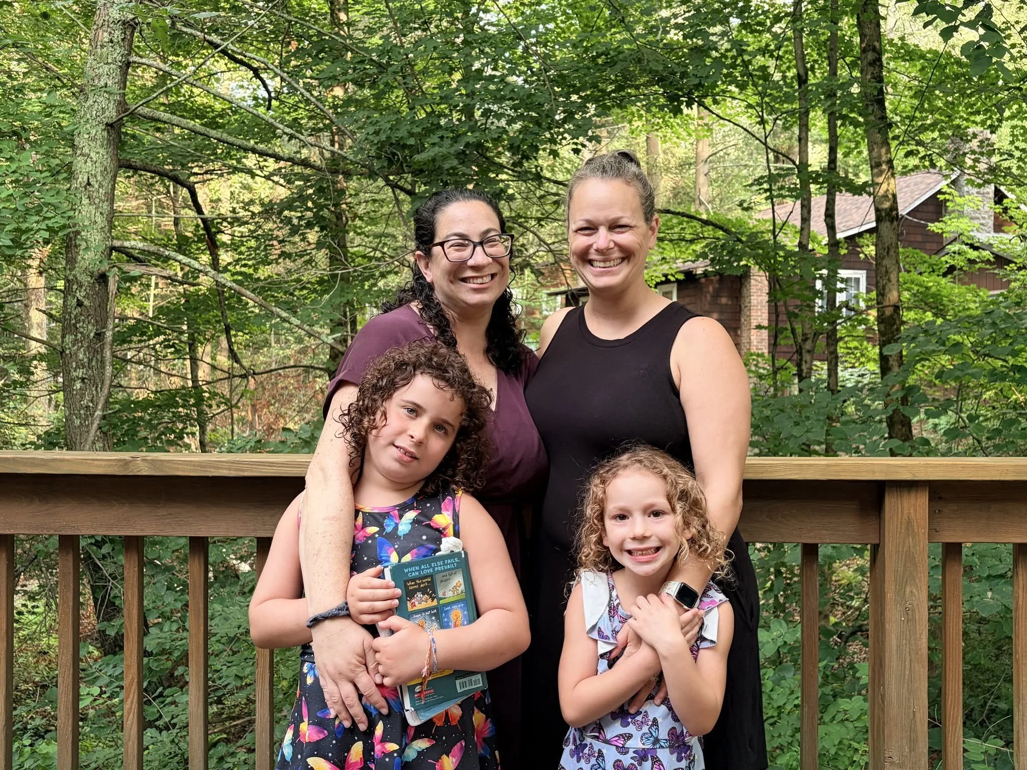 A group of five people, two adults and three children, smiling and standing on a wooden deck in a forested area with green foliage and trees in the background.