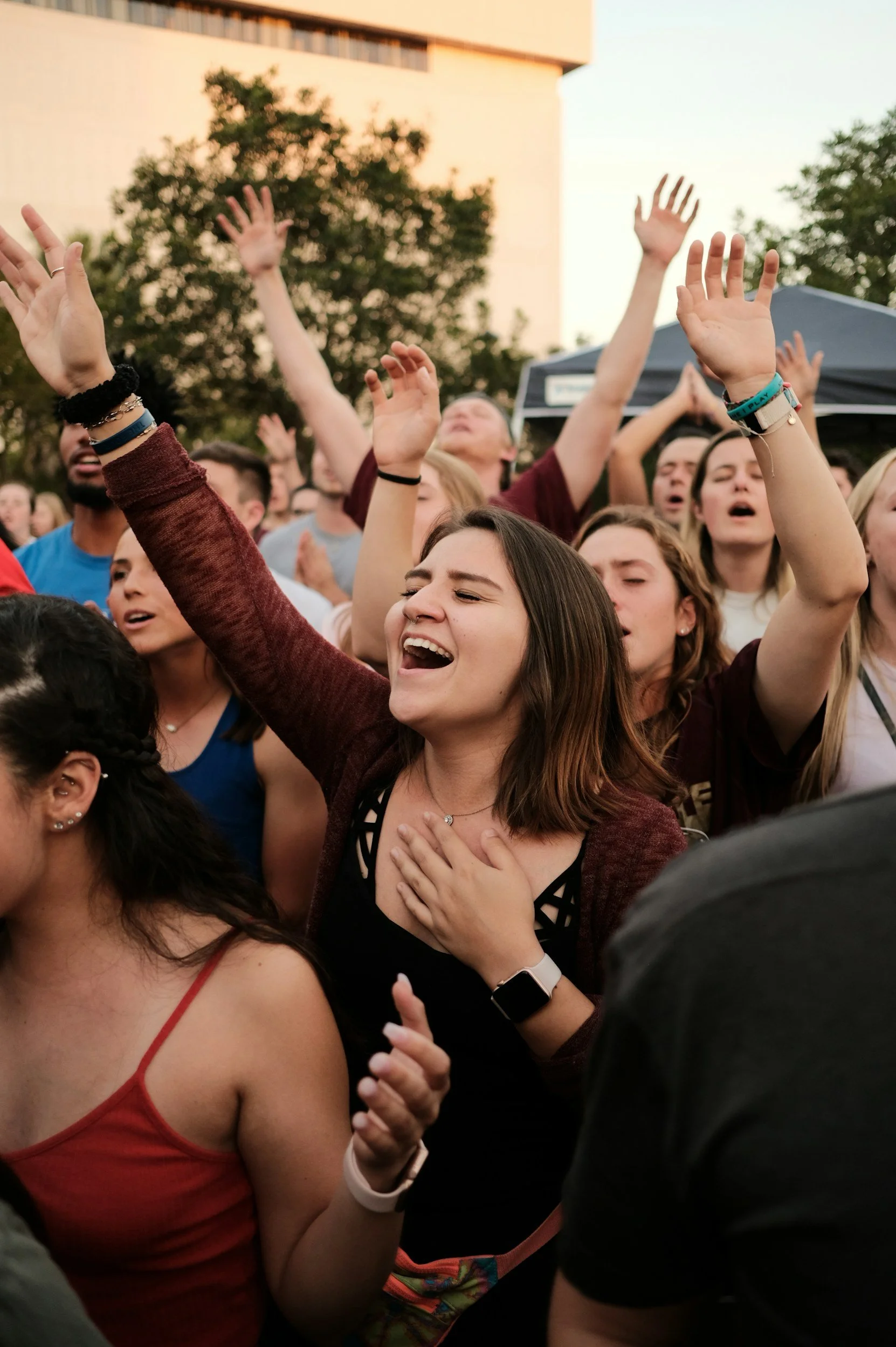 A crowd of people with eyes closed, some with guitars, singing or worshipping outdoors during sunset with trees and a building in the background.