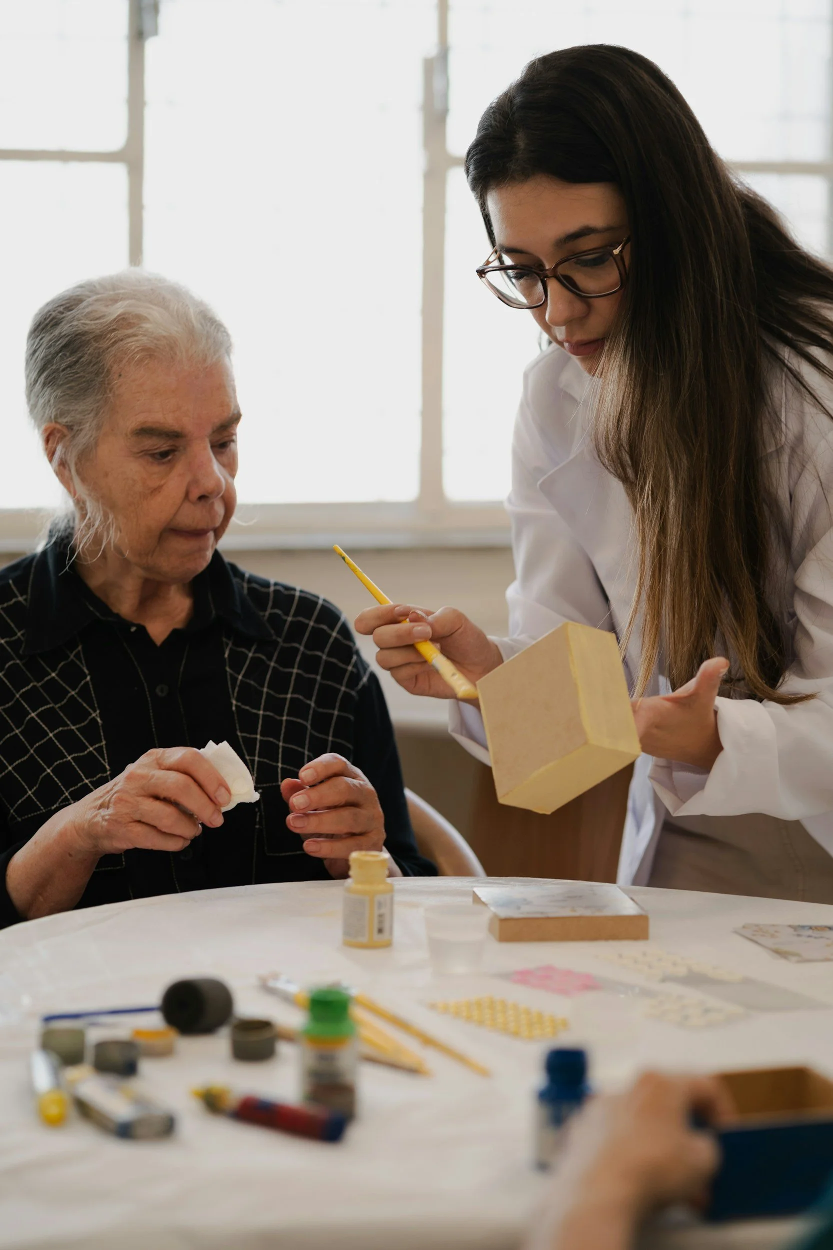A young woman in glasses helping an elderly woman with a craft project at a table. The table has craft supplies and small jars, and sunlight is streaming through the window.