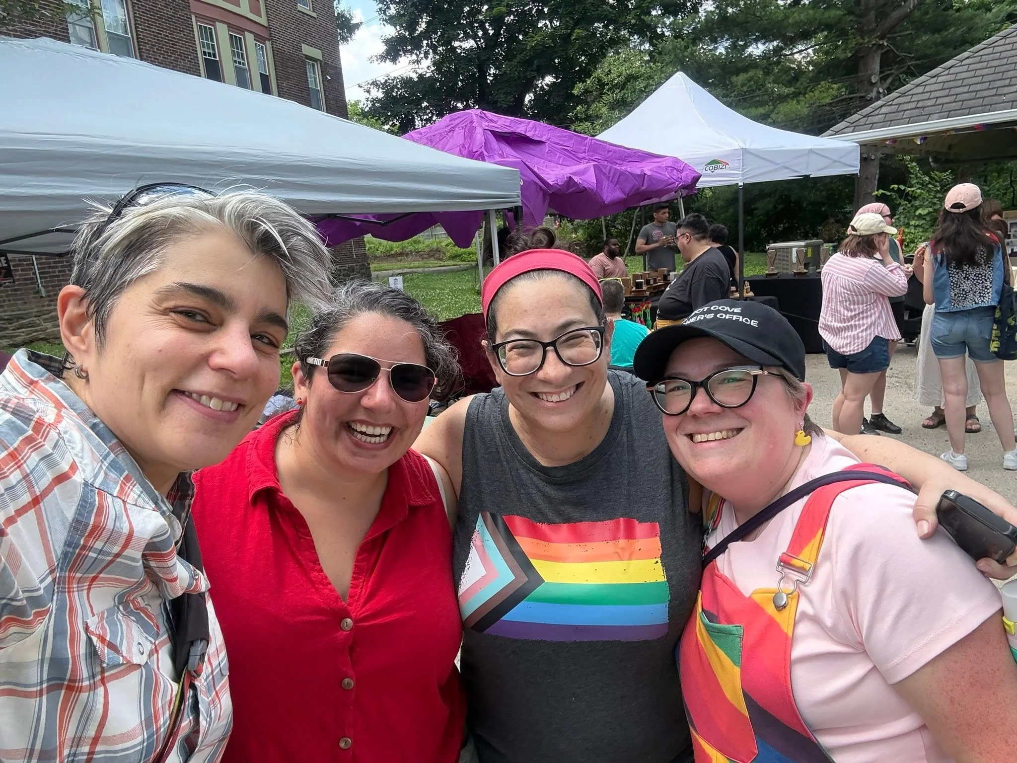 Group of smiling people taking a selfie at an outdoor event with tents and trees in the background.