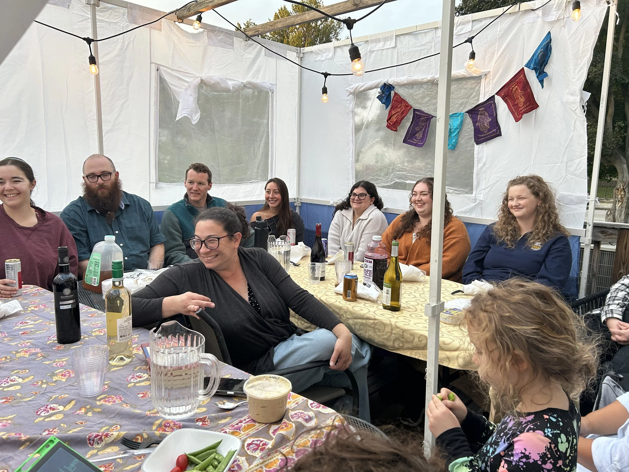 Group of people sitting at a table under a white canopy, smiling and enjoying a gathering or celebration, with food and drinks on the table, decorated with hanging string lights and colorful flags.