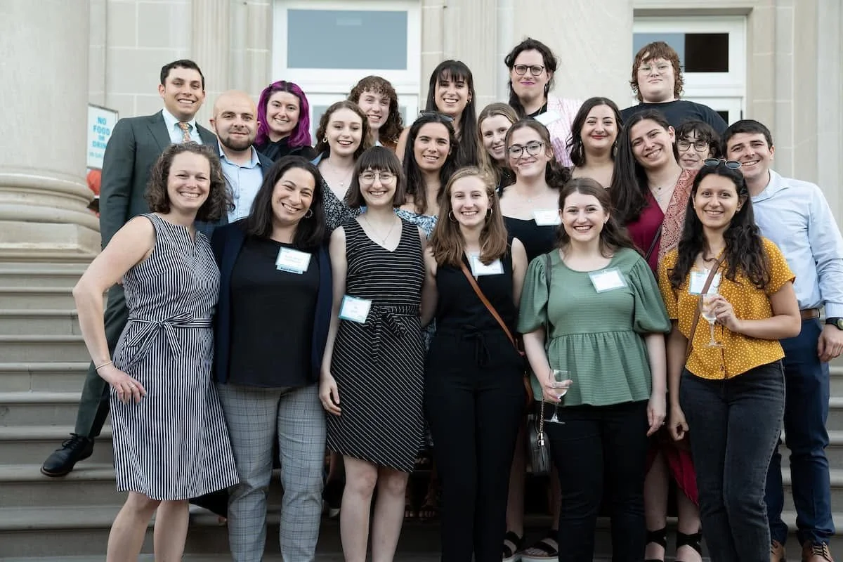 Group of 20 smiling young adults, mostly women, gathered on steps outside a building for a group photo at an event or social gathering.