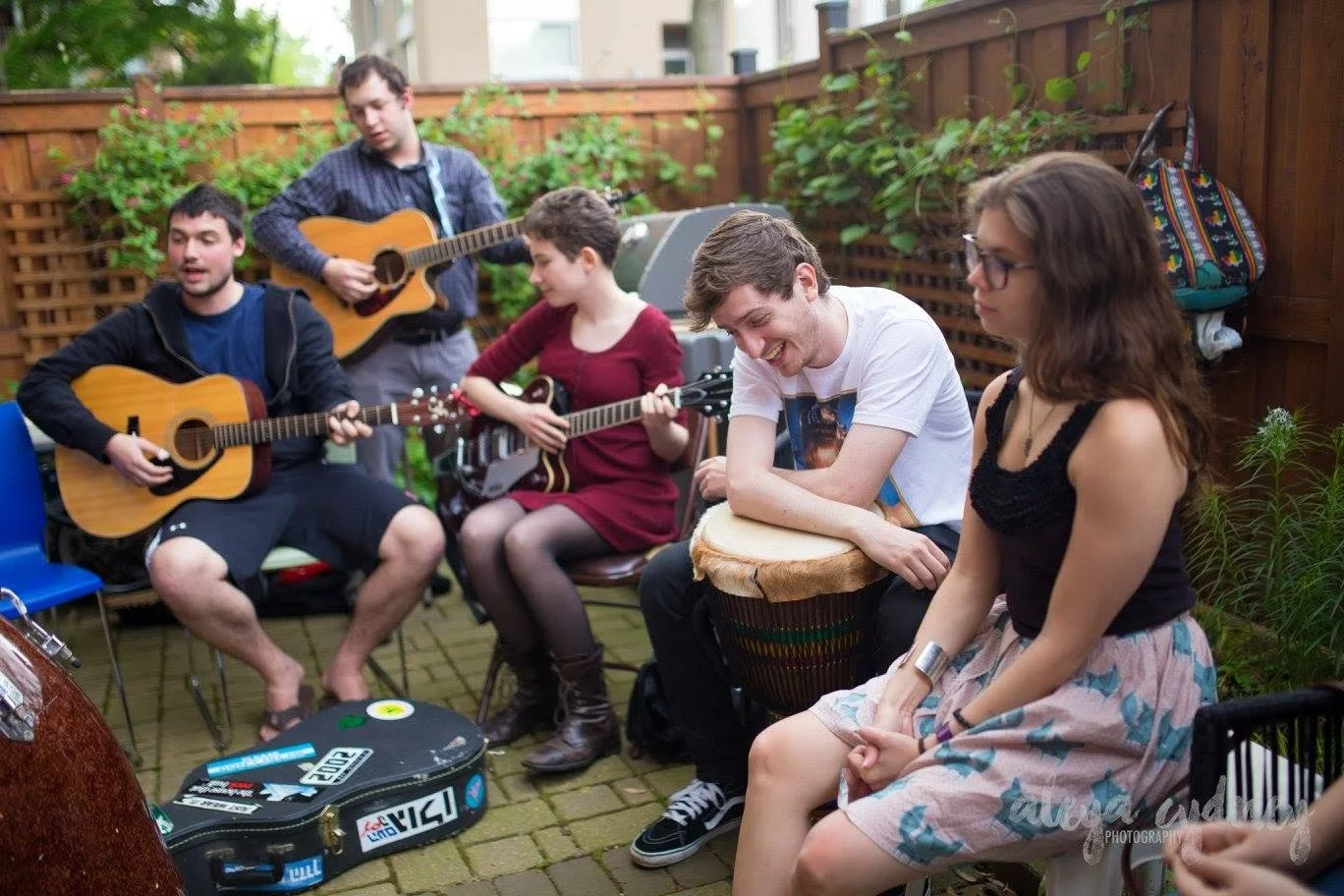 Group of five people playing guitars and a drum in an outdoor patio with wooden fencing and plants.