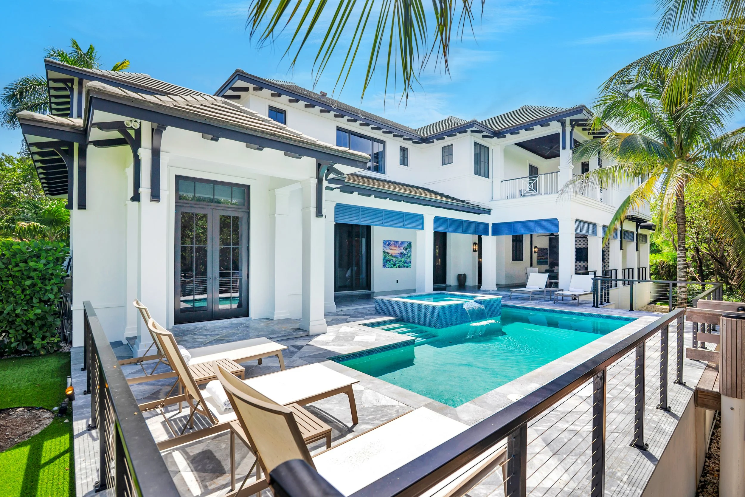 Modern house with a swimming pool, outdoor lounge chairs, surrounded by lush greenery and palm trees under a clear blue sky.