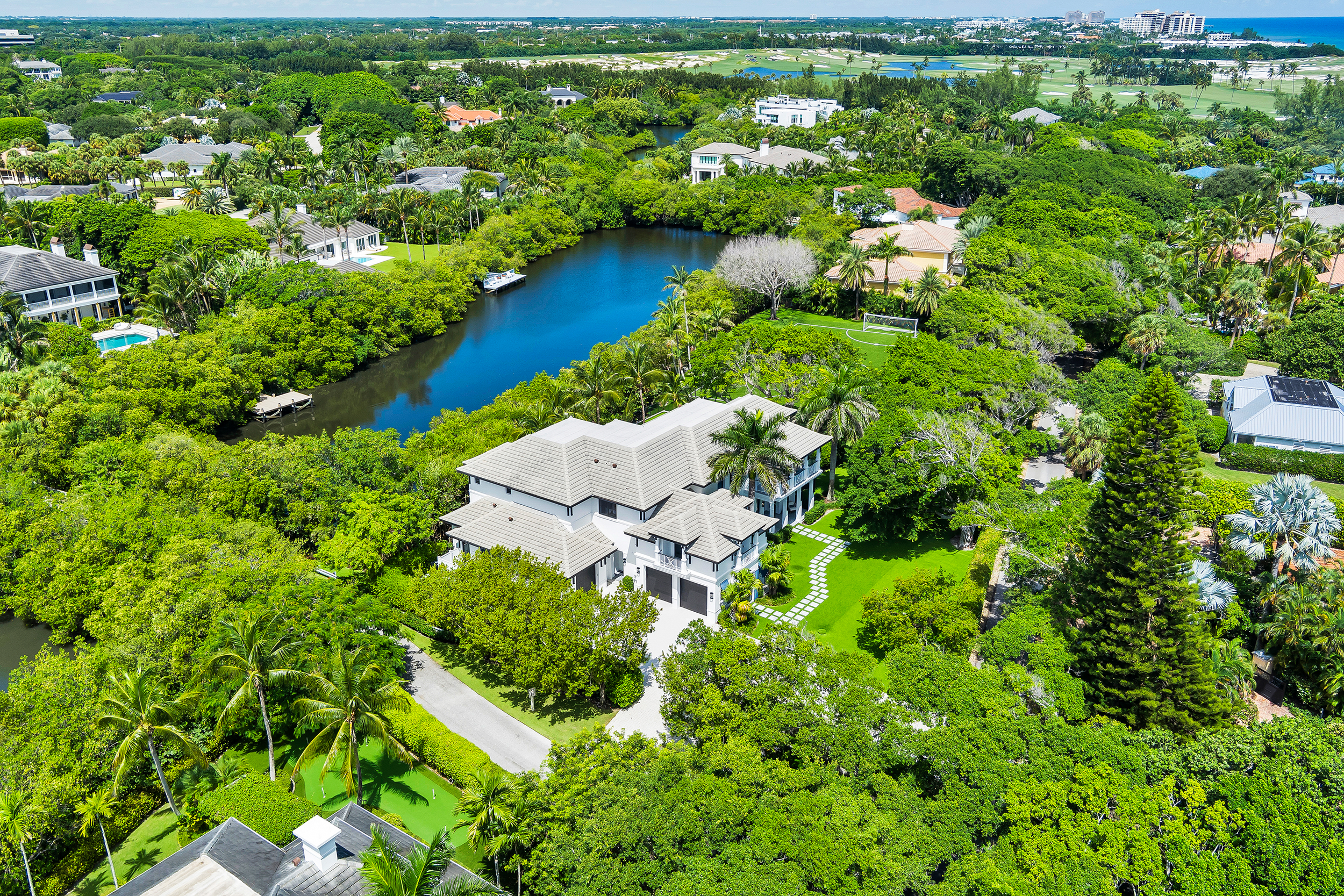 Aerial view of a lush tropical residential neighborhood with large houses, palm trees, a small lake, and a golf course in the background.