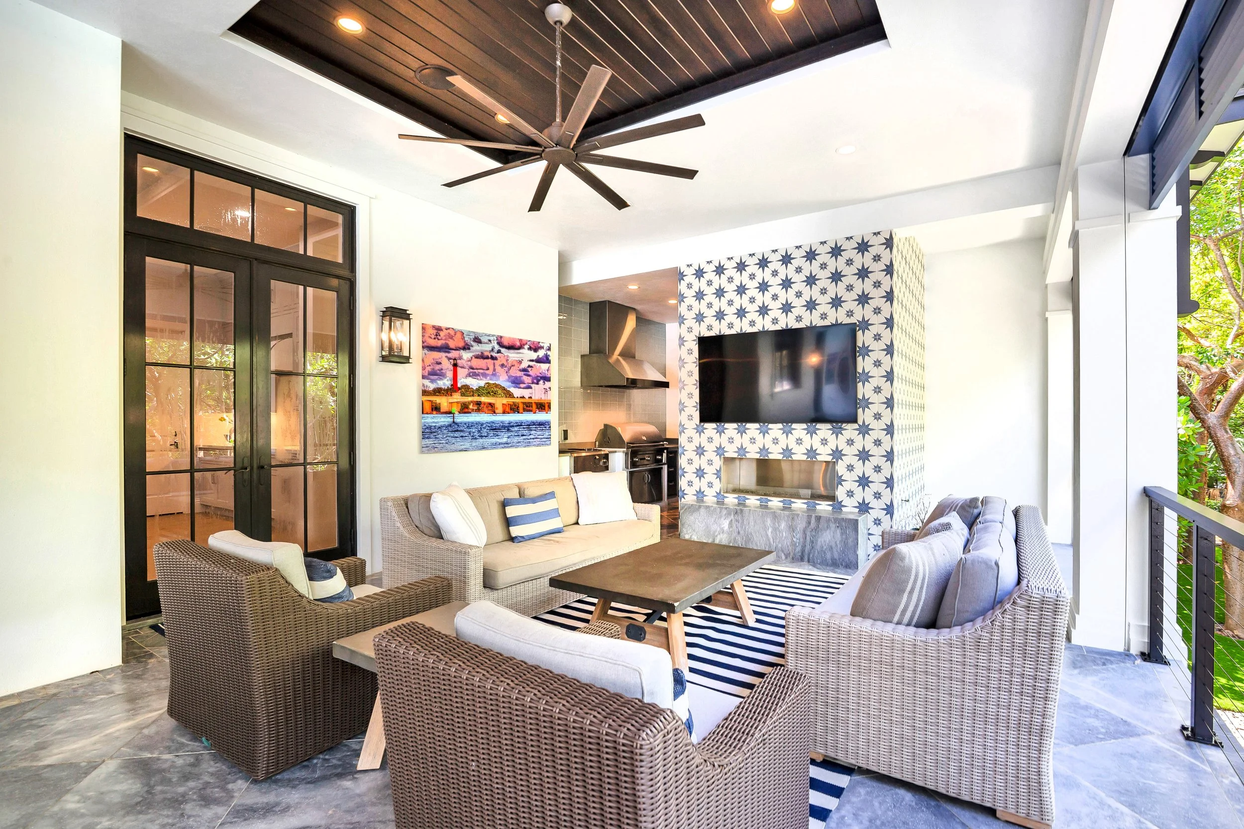 A spacious outdoor living area with wicker and upholstered chairs, a sofa, a coffee table, a flat-screen TV mounted on a blue patterned wall, a ceiling fan, and a view of a tree through an open balcony.