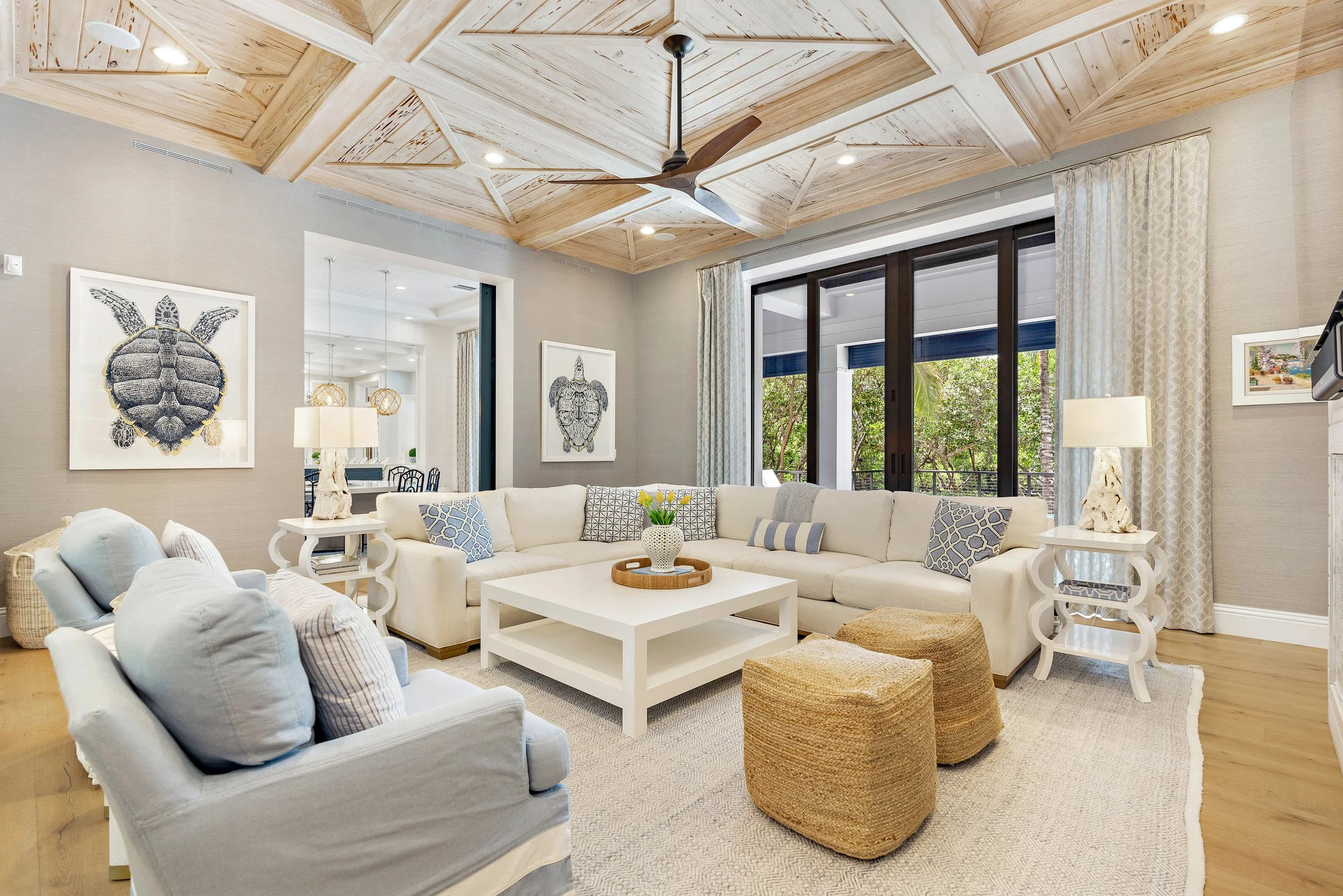 Living room with white sectional sofa, gray armchair, woven poufs, wooden coffee table with vase, beige curtains, and decorative turtle images on the wall. Wooden ceiling with fan and large glass doors showing outdoor greenery.