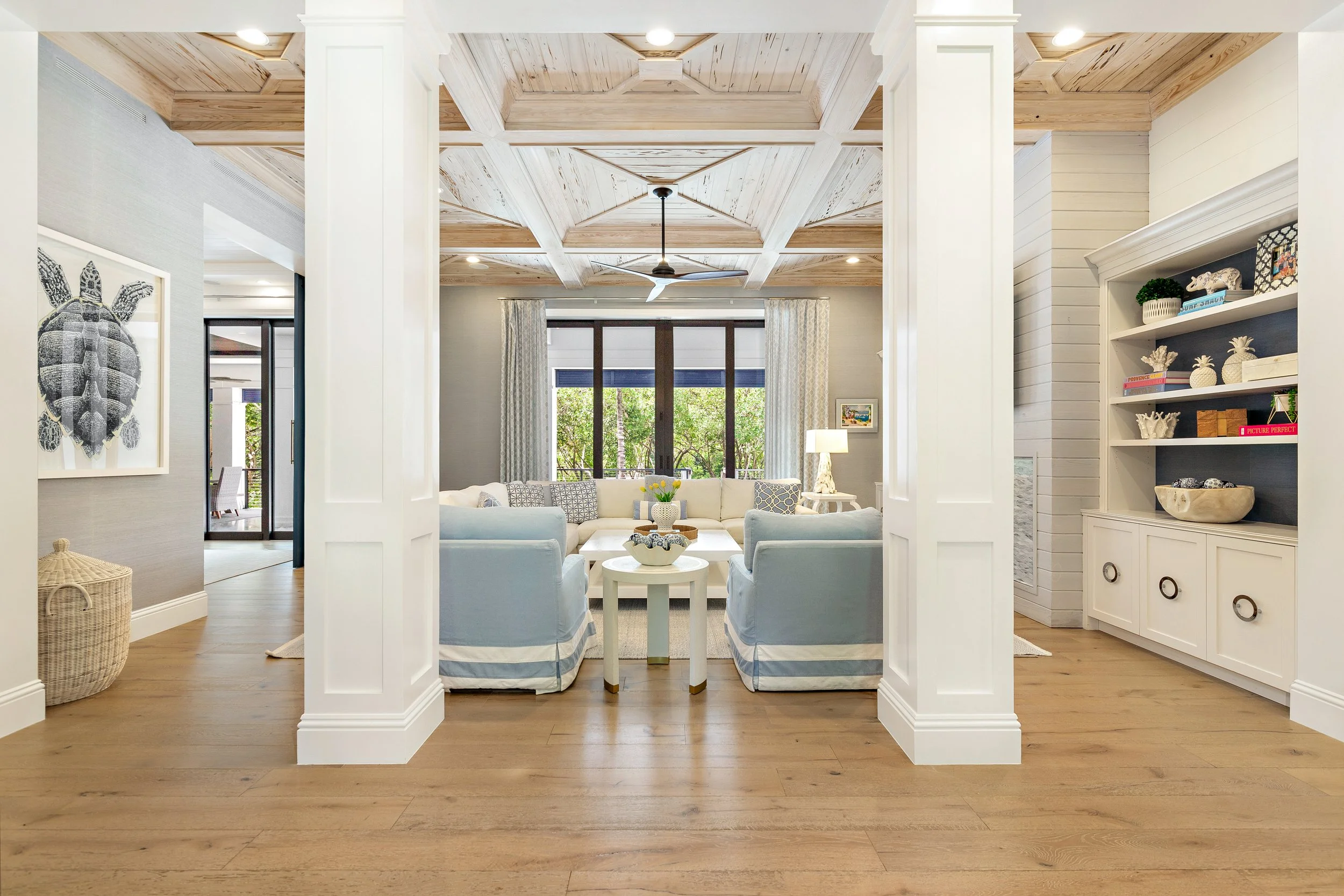 Living room with white couches, blue and white striped skirts, a white coffee table, large windows with curtains, and a bookshelf with decor. Wooden flooring and a ceiling with wood paneling and a ceiling fan.